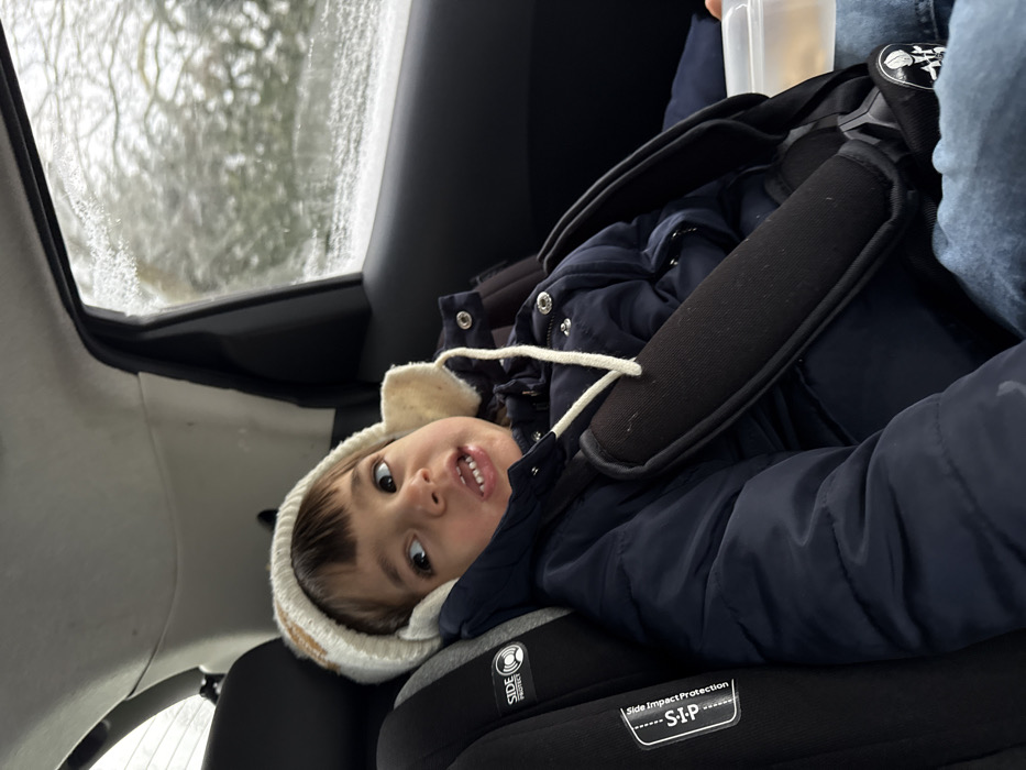 A young child, estimated to be between two and four years old, is depicted seated in the rear passenger compartment of a vehicle in Senningerberg, Luxembourg. The child is secured in a black child safety seat, marked with "Side Impact Protection" (S-I-P) branding, utilizing a multi-point harness system with black padded shoulder straps. The child wears a cream-colored knit winter hat with ear flaps and white securing strings, a dark blue puffy winter jacket, and light blue jeans. The child's gaze is directed upwards and to the right (from the viewer's perspective), with a slightly open mouth revealing front teeth. Partially visible on the child's right side, near their lap, is a clear plastic container holding light-colored contents. The vehicle's interior features a light-colored headliner and dark-colored door trim. Outside the vehicle's window, a blurred winter landscape is visible, characterized by bare tree branches indicative of cold weather and an overcast sky. The vehicle window itself exhibits minor frost or condensation along its upper and lower edges.