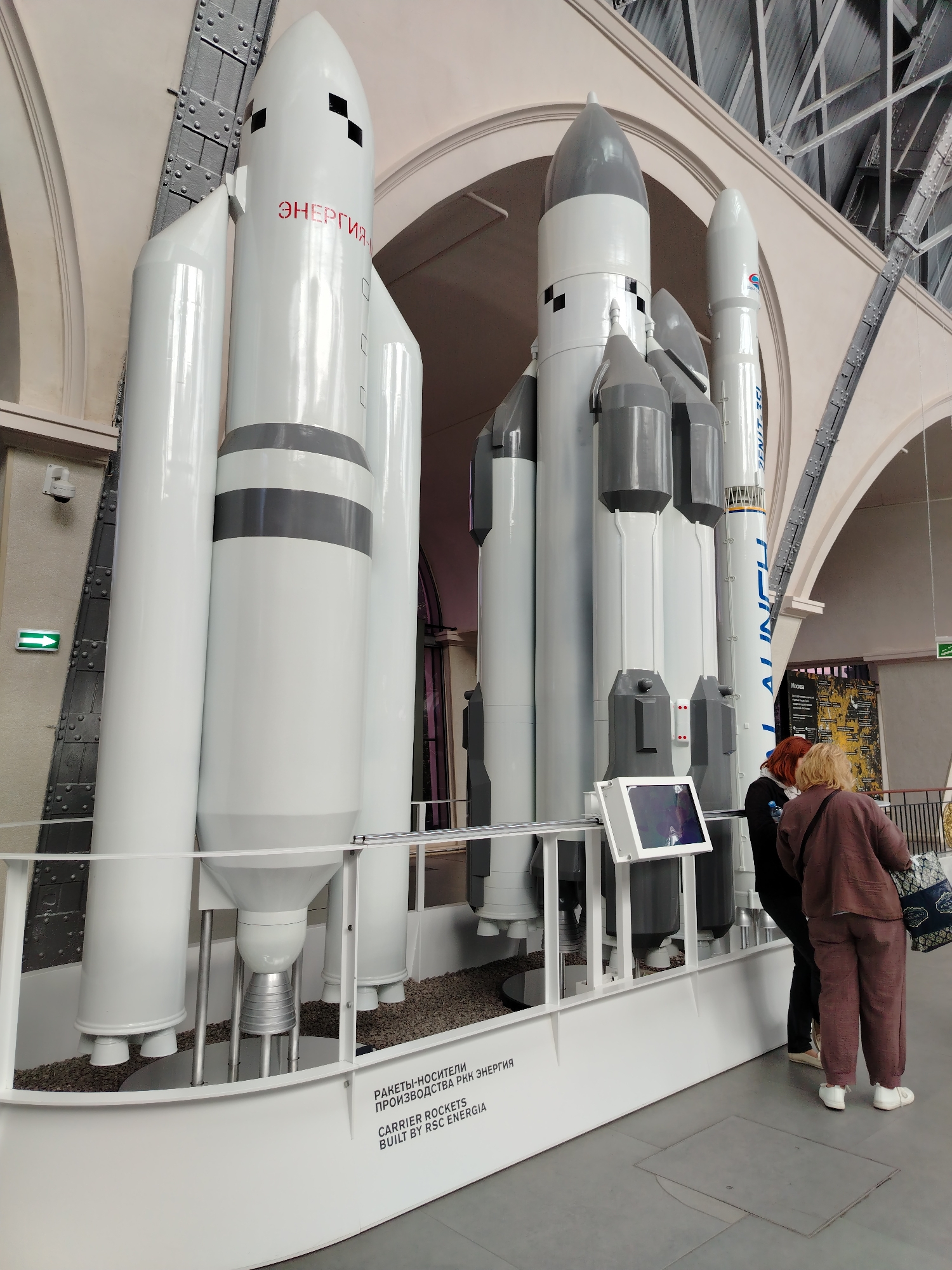 In a museum hall in Moscow, Russia, a large model of the Soviet-era Energia rocket is displayed. The rocket is white with grey accents and is accompanied by smaller rocket models. Two women are standing in front of the exhibit, looking at the display. The hall has high ceilings with exposed metal beams and arched architectural elements. The floor is a light gray concrete, and there is a railing around the exhibit. A green illuminated exit sign is visible on the left wall. The text on the exhibit reads "РАКЕТЫ-НОСИТЕЛИ ПРОИЗВОДСТВА РКК ЭНЕРГИЯ" and "CARRIER ROCKETS BUILT BY RSC ENERGIA".
