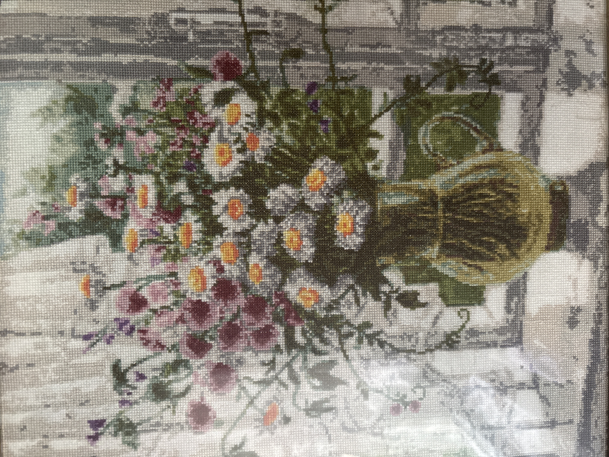 This image depicts a cross-stitch artwork of a floral arrangement in a vase. The artwork shows a bouquet of various flowers, including white daisies with orange centers and pink and deep red blossoms, situated in a golden-hued glass vase. The vase rests on a surface that appears to be a windowsill. Behind the flowers, a blurred background suggests a window with a view of greenery. The artwork is framed by what looks like a decorative border. There are no people visible in the image. The image is a close-up view of the cross-stitch itself, showing the texture of the threads and the detail of the stitching. There is no information or visual evidence within the image to suggest it is located in the city of Tomsk, Russia, or any specific geographic location.