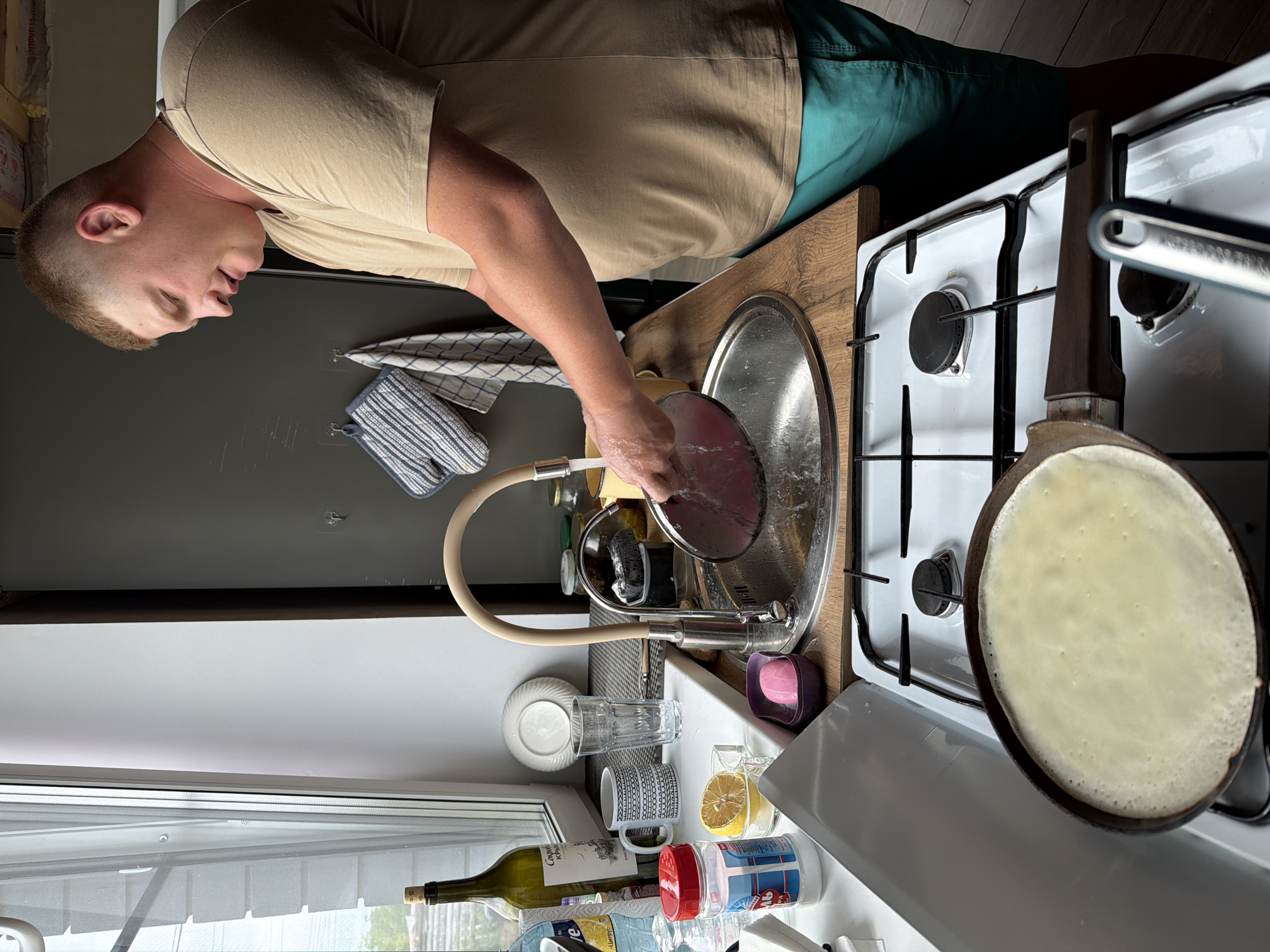 A man is washing a pot in a kitchen. The kitchen contains a sink, faucet, white gas stove with a pancake being cooked in a pan, a bottle of wine, and various other kitchen items. The man is wearing a tan t-shirt and green shorts. A window is visible in the lower left corner of the image. The location is Chaikovsky, Russia.