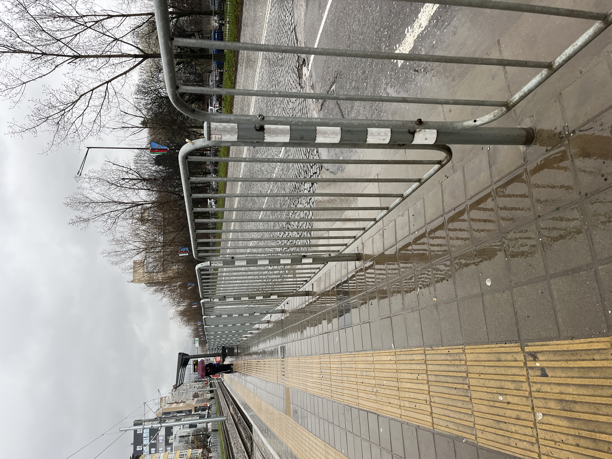 A wet, outdoor scene shows a train platform next to a street. A grey metal barrier with white stripes is adjacent to the street. A raised, yellow, tactile paving strip runs along the edge of the train platform. A person with a red umbrella stands further down the platform. Overhead electrical lines are visible, as are buildings across the tracks. The photo was taken on a cloudy day. The image is taken in Sofia, Bulgaria.