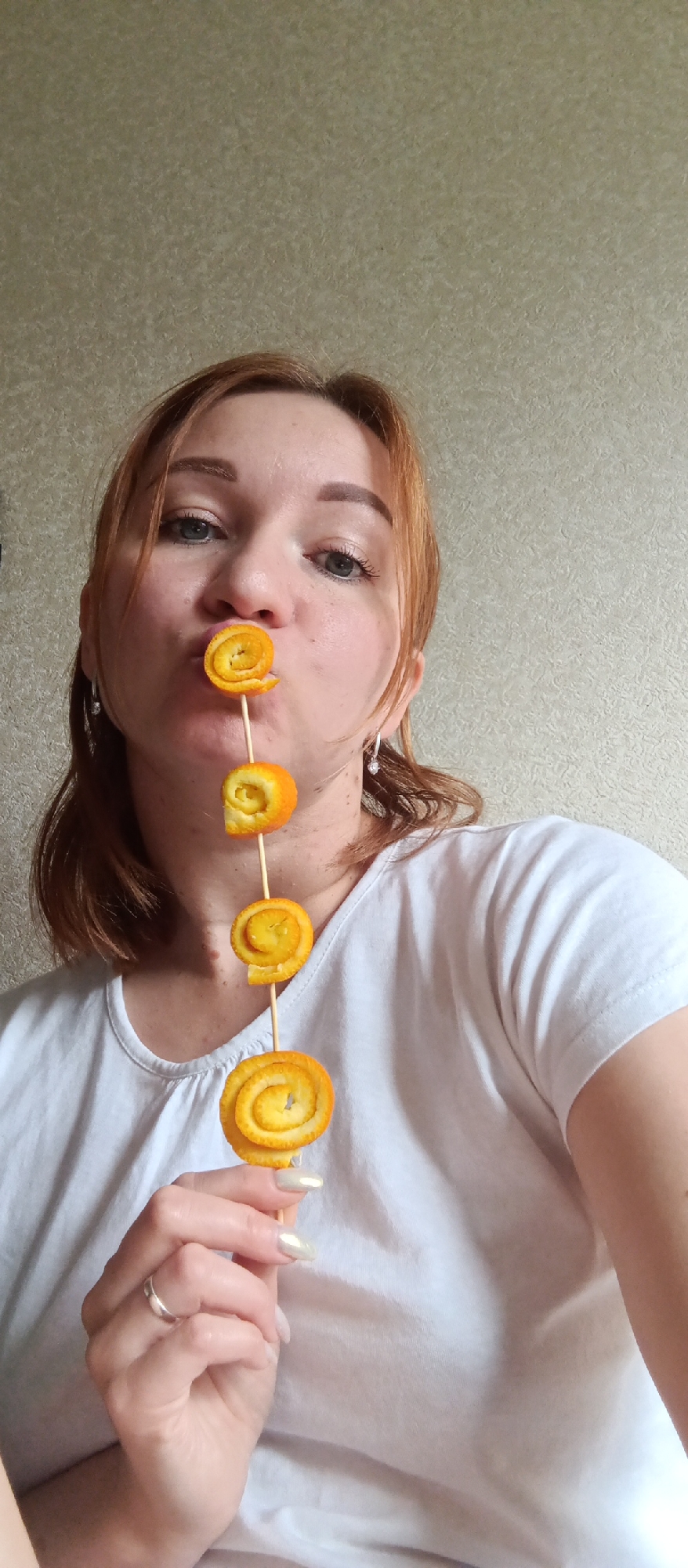 A woman with red hair and fair skin holds a stick with four orange peel spirals skewered on it. The topmost spiral is held in her mouth. She wears a white t-shirt, earrings, and a ring on her left hand. The background is a plain, light-colored wall. The location is Unknown, Unknown.