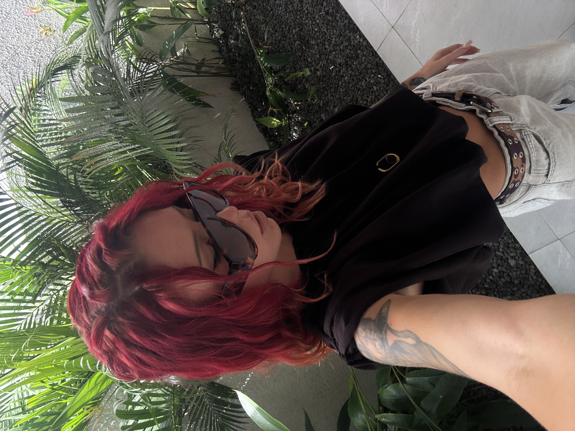 A woman with red wavy hair and sunglasses is photographed outdoors. She is wearing a black top and light-colored jeans with a spotted belt. Part of a tattoo is visible on her left arm. The background includes tropical foliage, dark gravel, and light-colored tiles. The location is North Kuta, Indonesia.