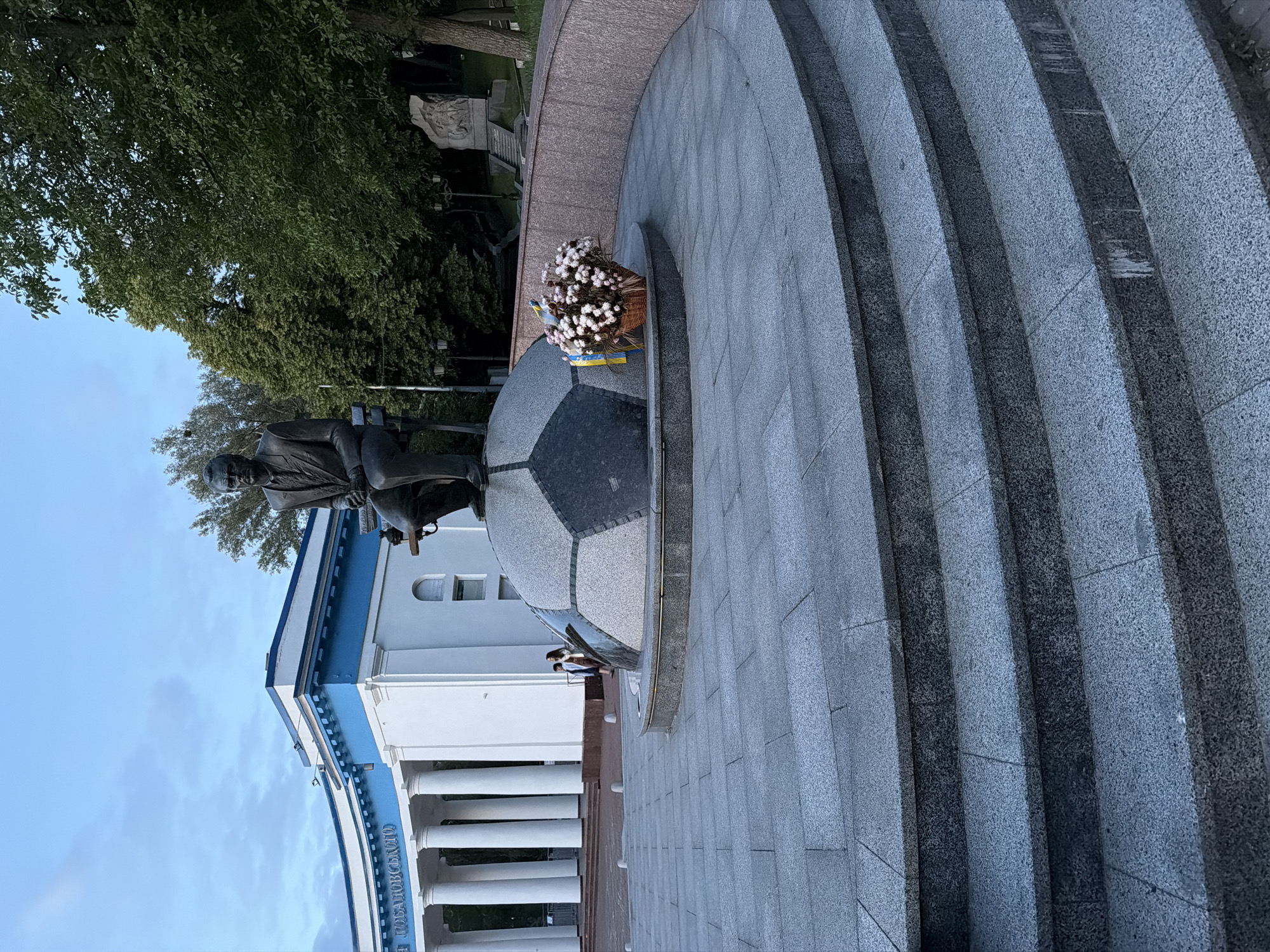 The image depicts a bronze statue of a man seated on a bench atop a large, round pedestal designed to resemble a soccer ball. The statue is located in a public square in Kyiv, Ukraine, specifically in front of the Lobanovsky Dynamo Stadium. To the left of the statue, the white and blue facade of the stadium with its prominent columns is visible. A basket of white flowers with a Ukrainian flag draped over it sits on the pedestal, to the right of the statue. In the background, lush green trees line a pathway leading to a sculptural monument. A few individuals are walking in the middle ground, near the stadium entrance. The foreground features a curved set of wide, grey stone steps. The sky is overcast with a pale blue hue.