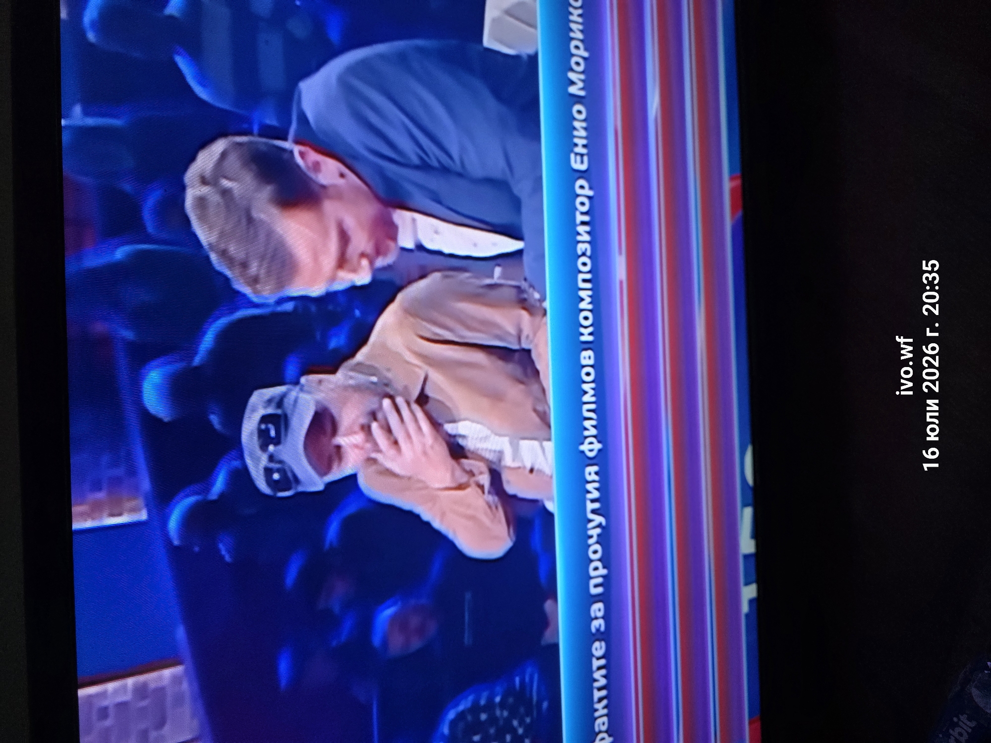 The image shows a close-up, slightly blurry, full-frame shot of a television screen. The screen displays two men seated at a table, with other people blurred in the background. The man on the left is wearing a light brown jacket over a white shirt and a white baseball cap with dark sunglasses. He is covering his mouth with his hand, appearing to be in distress or surprise. The man on the right is wearing a dark blue suit with a white shirt and a light-colored tie. He is looking down at his hands, which are resting on the table in front of him. A blue banner with white text is visible across the bottom of the screen, partially obscuring the lower bodies of the men. The text on the banner appears to be in Cyrillic script. Below the television screen, in the lower portion of the image, there is blackness and some indistinct text, including "ivo.wf" and "16 юли 2026 г. 20:35". The overall scene suggests a televised program or broadcast. No specific location in Chal, Bulgaria is discernible from the image content.