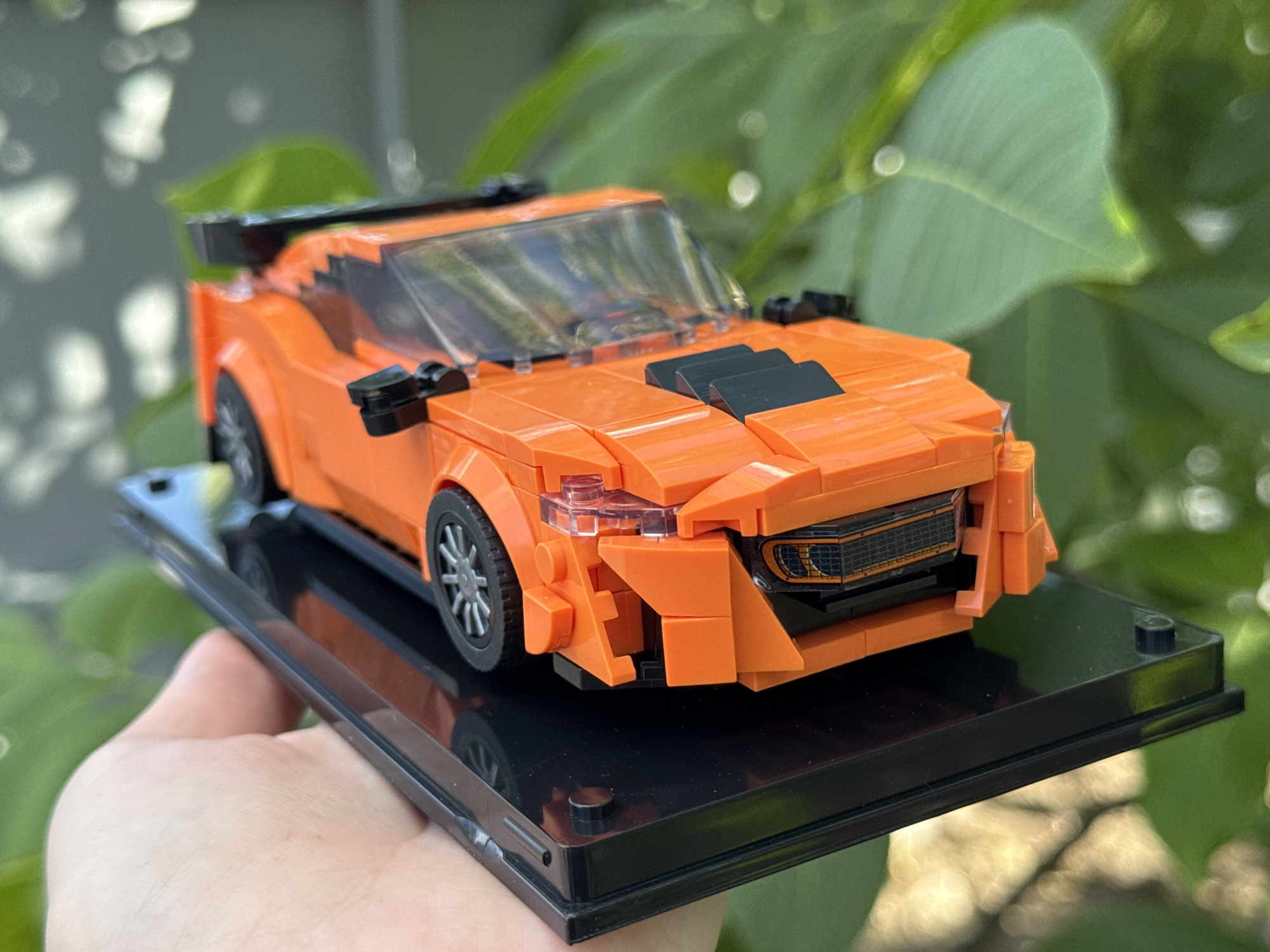 An orange Lego car model with black accents, including a spoiler and hood scoops, sits on a black display base. The car has intricate detailing, with clear Lego pieces used for the headlights and windshield. The display base is being held by a person's hand, partially visible at the bottom left of the frame. The background consists of blurred green foliage, suggesting an outdoor setting.