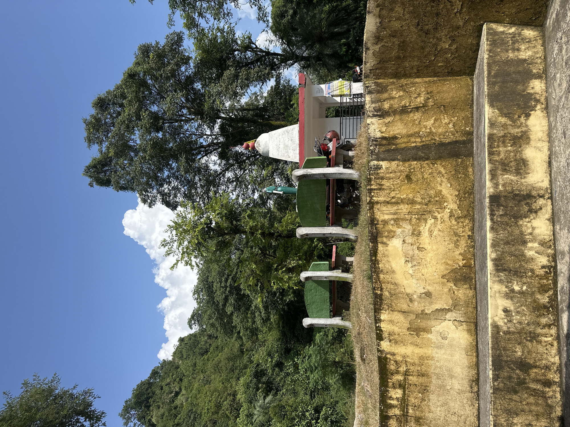 This is a photograph of a serene outdoor scene, likely in Kohla, India, given the context. In the foreground, a weathered, yellow-stained concrete wall forms a tiered structure, with two visible levels. Beyond this wall, a patch of dry grass supports two park benches with green seating and grey stone armrests. To the right of the benches, a small white temple with a red-tiled roof stands prominently. The temple features a conical finial at its peak, topped with red spheres. A flag, partially visible behind the temple, appears to be green and white.
Several motorcycles are parked near the temple and benches. A green statue, possibly of a deity or person, is positioned to the left of the temple. The background is dominated by lush, dense green foliage and tall trees, characteristic of a hilly or forested region. The sky above is a vibrant blue, dotted with fluffy white clouds. The overall atmosphere is peaceful and natural, suggesting a place of religious or recreational significance amidst nature.