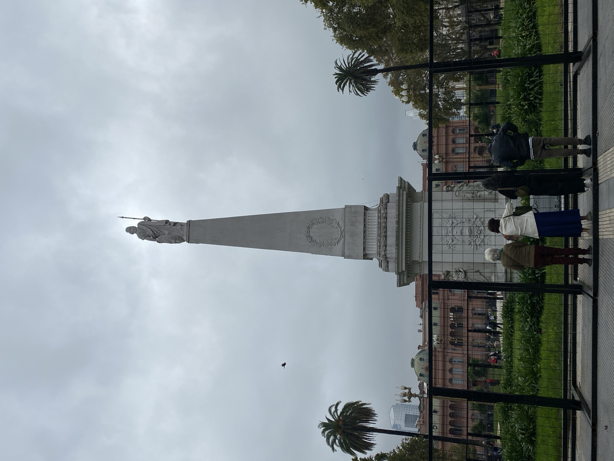 The image depicts a public square or park scene on an overcast day, dominated by a tall, light-colored stone monument. The monument, resembling an obelisk or column, features a statue of a figure holding a spear at its top, positioned towards the left of the frame. A circular wreath carving is visible on the monument's base.
In the background, to the right of the monument, a distinctive multi-story building with a reddish-pink facade, classical architectural elements, and prominent domes on its roof is visible. This building is partially obscured by a black metal fence that stretches across the mid-ground. Several individuals are present, with at least four people seen from behind in the foreground, standing near the fence and seemingly observing the monument or the pink building.
The scene includes natural elements such as two prominent palm trees – one in the upper right and another in the lower right – and other green foliage and trees. The sky is largely covered by light grey clouds, indicating an overcast daytime. A small dark bird is captured in flight against the cloudy sky. The foreground shows a paved area where the observers stand, separated from the monument and pink building by the metal fence. The pink building's unique appearance strongly suggests the location is the Casa Rosada in Buenos Aires, Argentina. No discernible text is visible on the monument or buildings.