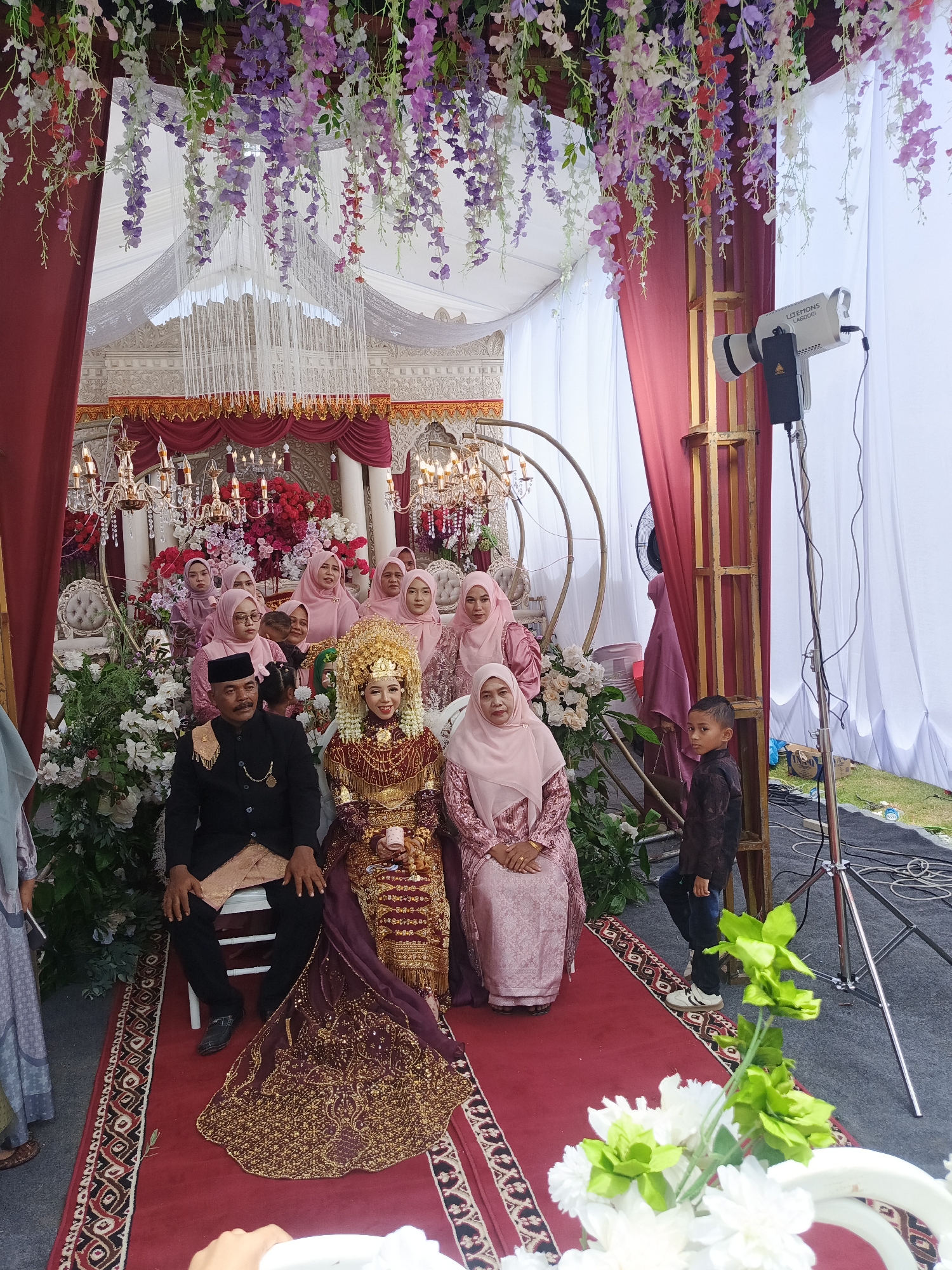 This is a full shot image of an outdoor wedding ceremony. A man and woman, dressed in traditional attire, are seated on a red carpet in front of a backdrop of white fabric and draped red curtains. The woman is adorned in a gold headdress and a richly embellished red and gold dress. The man is wearing a black suit with a gold sash. Behind them, a group of women wearing pink headscarves and dresses are seated, observing the ceremony. The scene is decorated with abundant floral arrangements in shades of pink, purple, and white, as well as ornate chandeliers. A young boy in a dark shirt and jeans stands to the right, and a lighting rig is visible in the background. The event appears to be taking place in Meuliek, Indonesia.