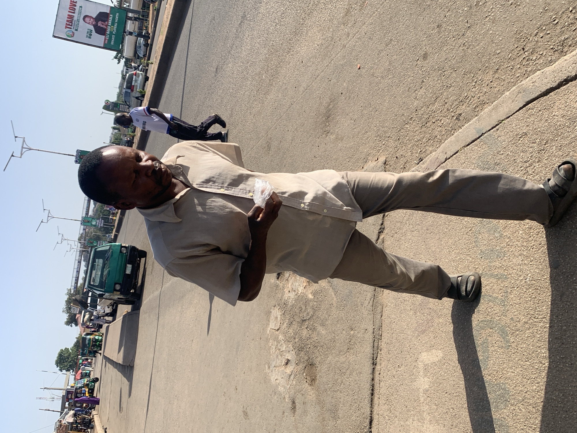An adult male stands on a paved road, holding a small plastic bag. He wears a light-colored button-up shirt and dark pants with dark sandals. The road is lined with sidewalks, and in the background is a billboard for "TEAM LOVE" and a candidate for Senate. Further in the background, there are vehicles including a green van and auto-rickshaws. Another person is visible lying on the side of the road in the distance. The scene appears to be set in an urban environment, near the city of Gura Top, Nigeria, given the objects and details present.