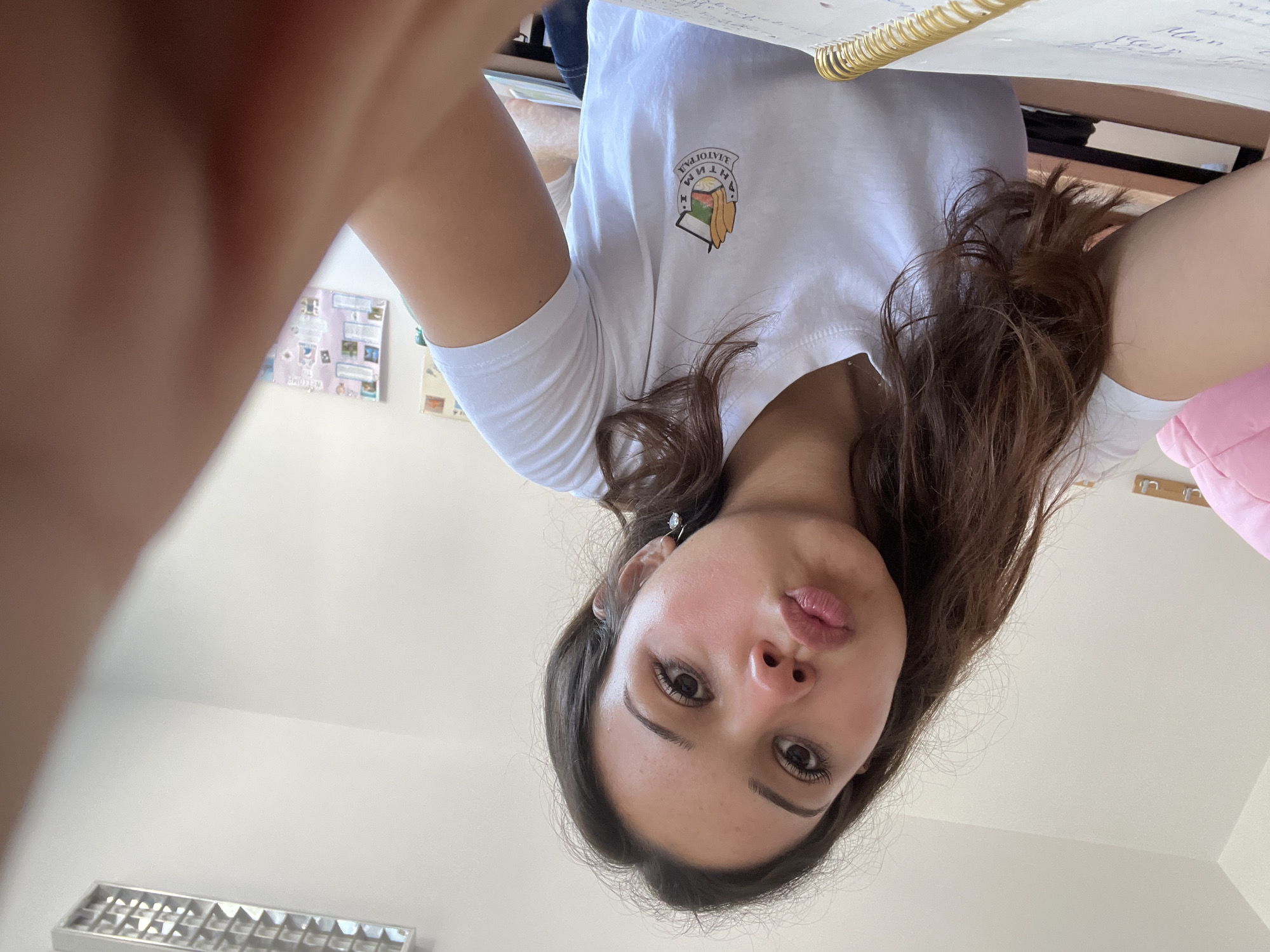 In a brightly lit room, a young woman with long, dark, wavy hair is captured in a selfie taken from a low angle. She is wearing a white t-shirt with a circular emblem on the left chest that features a shield-like design with what appears to be a landscape and the text "ЗЛАТОГРАД" (Zlatograd) in Cyrillic script. Her lips are slightly pursed, and she is looking directly at the camera. A small, sparkling earring is visible on her right ear. The background includes a white ceiling with fluorescent lighting, a portion of a pink cushion, and a corkboard with various pinned items and papers on the wall. A notebook with a gold spiral binding and handwritten notes is in the foreground. The overall environment suggests an indoor setting, possibly a classroom or a study area in Zlatograd, Bulgaria.