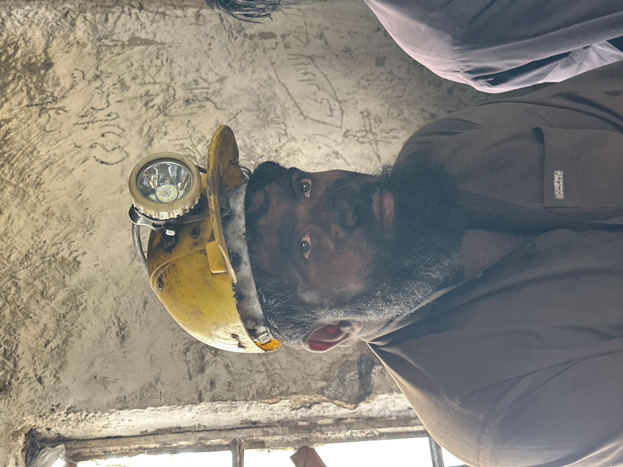 In Quetta, Pakistan, a miner, his face and beard smudged with coal dust, looks to his right. He wears a yellow hard hat equipped with a headlamp, a brown shirt with a small "Lucky" label on the chest pocket, and has a dark beard. Behind him, graffiti is visible on a rough, textured wall. To the right of the frame, another person in a light purple shirt is partially visible. The setting appears to be an interior space, possibly a mine entrance or a related structure.
