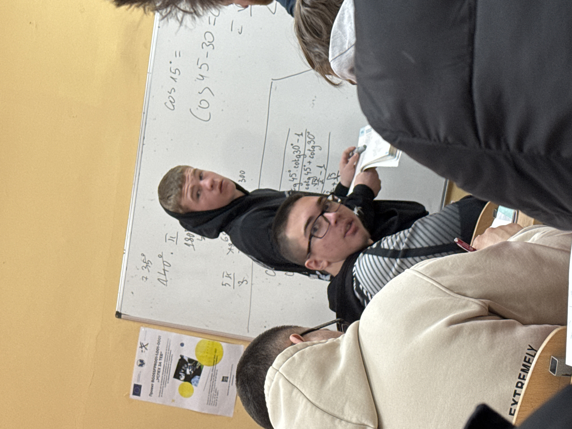 Here is a description of the image:
**Scene:** A classroom setting is shown with a group of students around a whiteboard. The location appears to be a school, possibly in Varna, Bulgaria, given the poster on the wall includes Bulgarian text.
**People:** Several young people are present, likely high school or college students. One person is writing on the whiteboard, while others are looking at a notebook or textbook.
**Subjects:** The primary subject is a mathematics lesson being taught in the classroom. Equations are written on the whiteboard involving trigonometric functions such as cosine and cotangent.
**Objects:** The scene includes a whiteboard covered with mathematical formulas, notebooks, and pens, typical of a classroom.
**Actions:** The actions involve teaching, writing, and studying mathematical equations.
**Interactions:** Students are gathered around the whiteboard, suggesting collaboration or a teaching session.
**Environment:** The environment is a brightly lit classroom with yellow walls.