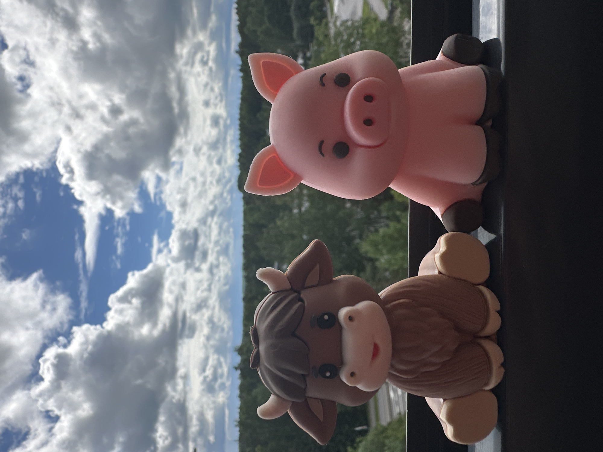 This media file features two small, cartoonish figurines: a pink pig and a brown cow, possibly made of fondant or a similar material. The figurines are positioned on a dark, flat surface, likely a balcony railing or windowsill, with their bottoms facing the camera.
Behind the figurines, the background is divided into two distinct areas. The left side shows a bright blue sky filled with large, fluffy white and gray clouds. The right side of the background is a blurred landscape of green trees and what appears to be distant foliage, suggesting an outdoor setting.
The pig, on top, is depicted with its characteristic pink ears, snout, and small, dark eyes and mouth. It is facing forward, with its body slightly turned to the side. The cow, positioned below the pig, has a brown body with lighter brown detailing that mimics fur or knitting, along with a white muzzle, dark eyes, and small horns. Both figurines appear to be standing or sitting.
The lighting suggests it is daytime, possibly late morning or afternoon, given the brightness of the sky. The presence of clouds indicates partly cloudy weather. The overall scene conveys a playful and cheerful mood, emphasized by the whimsical nature of the figurines. There is no visible text in the image.
