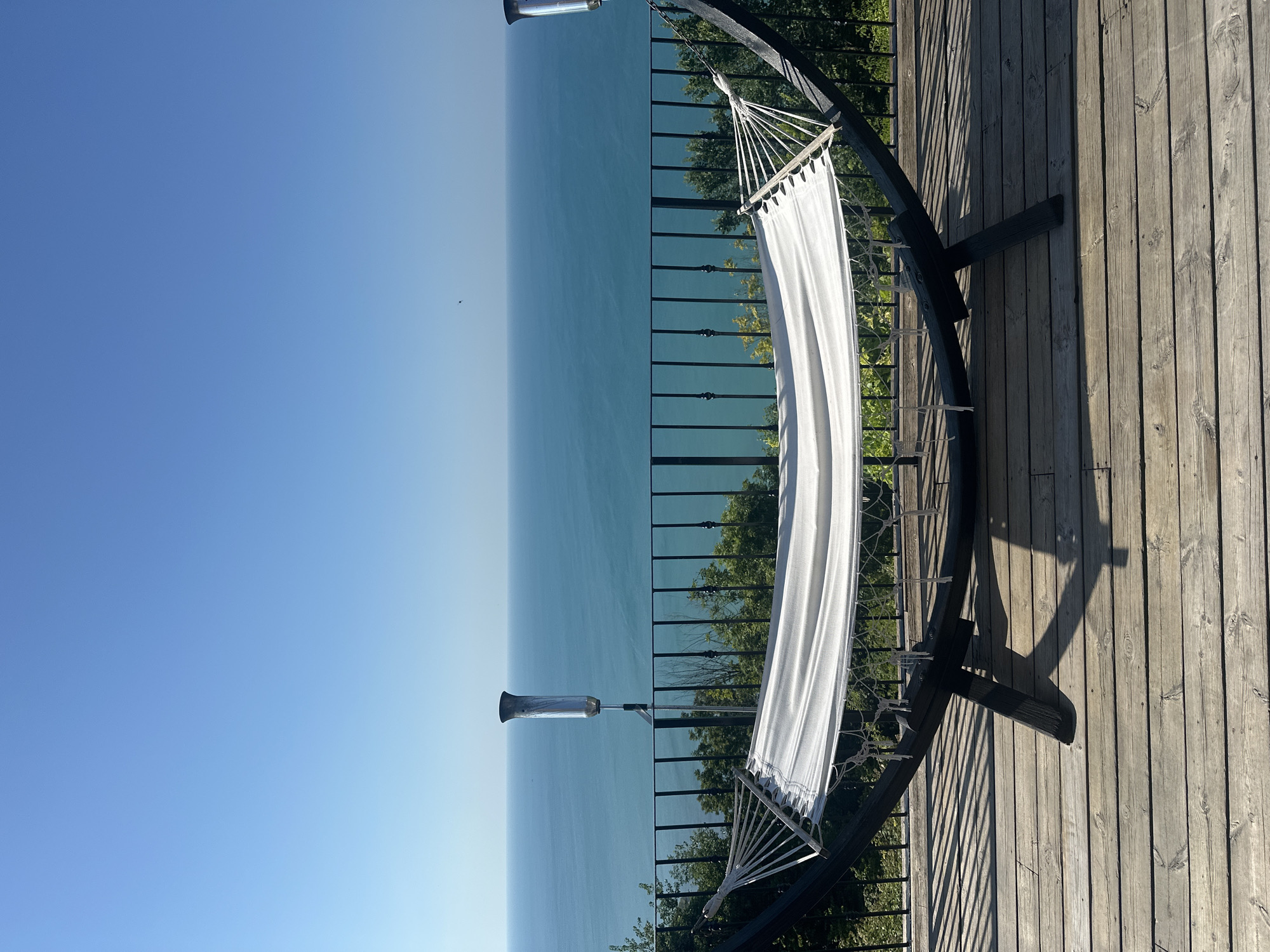 A hammock on a wooden deck overlooks a vast, calm blue ocean under a clear sky. The hammock is white with a dark frame, and its ropes are clearly visible. A black metal railing separates the deck from the water, with green trees visible below the railing. Two cylindrical lamps, one on each side of the frame, are attached to posts behind the railing. The wooden deck boards show a pattern of sunlight and shadow.