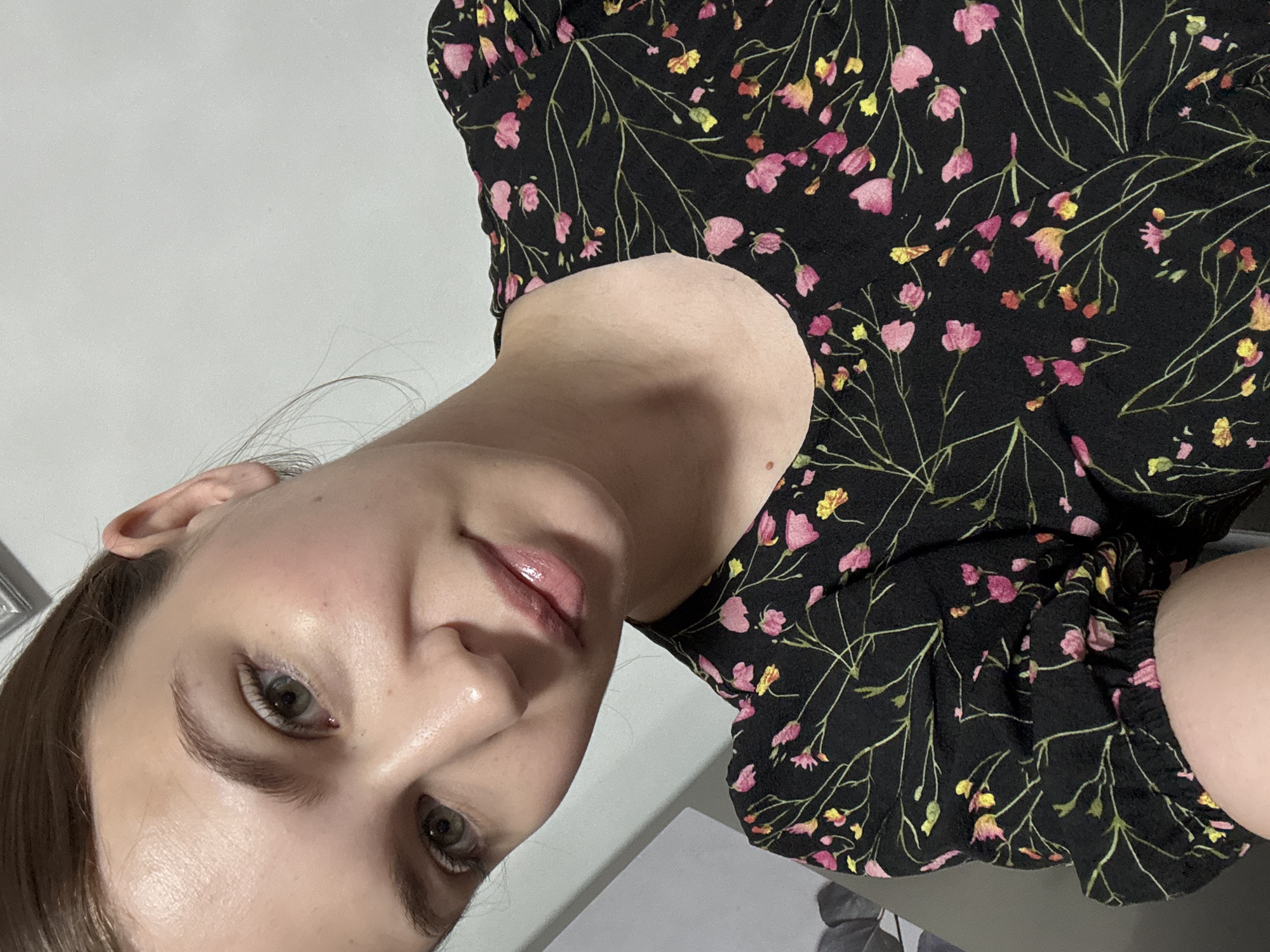 A selfie of a young woman taken indoors against a plain white wall. She has light skin, fair hair pulled back, and light green eyes. She is wearing a black dress with a floral pattern of pink and yellow flowers. Her head is tilted, and she is looking towards the camera. The lighting is soft, highlighting the natural glow of her skin and the glossiness of her lips. The background is a simple, unadorned wall, suggesting a domestic setting. There is no visible indication of the location being Chaikovsky, Russia.