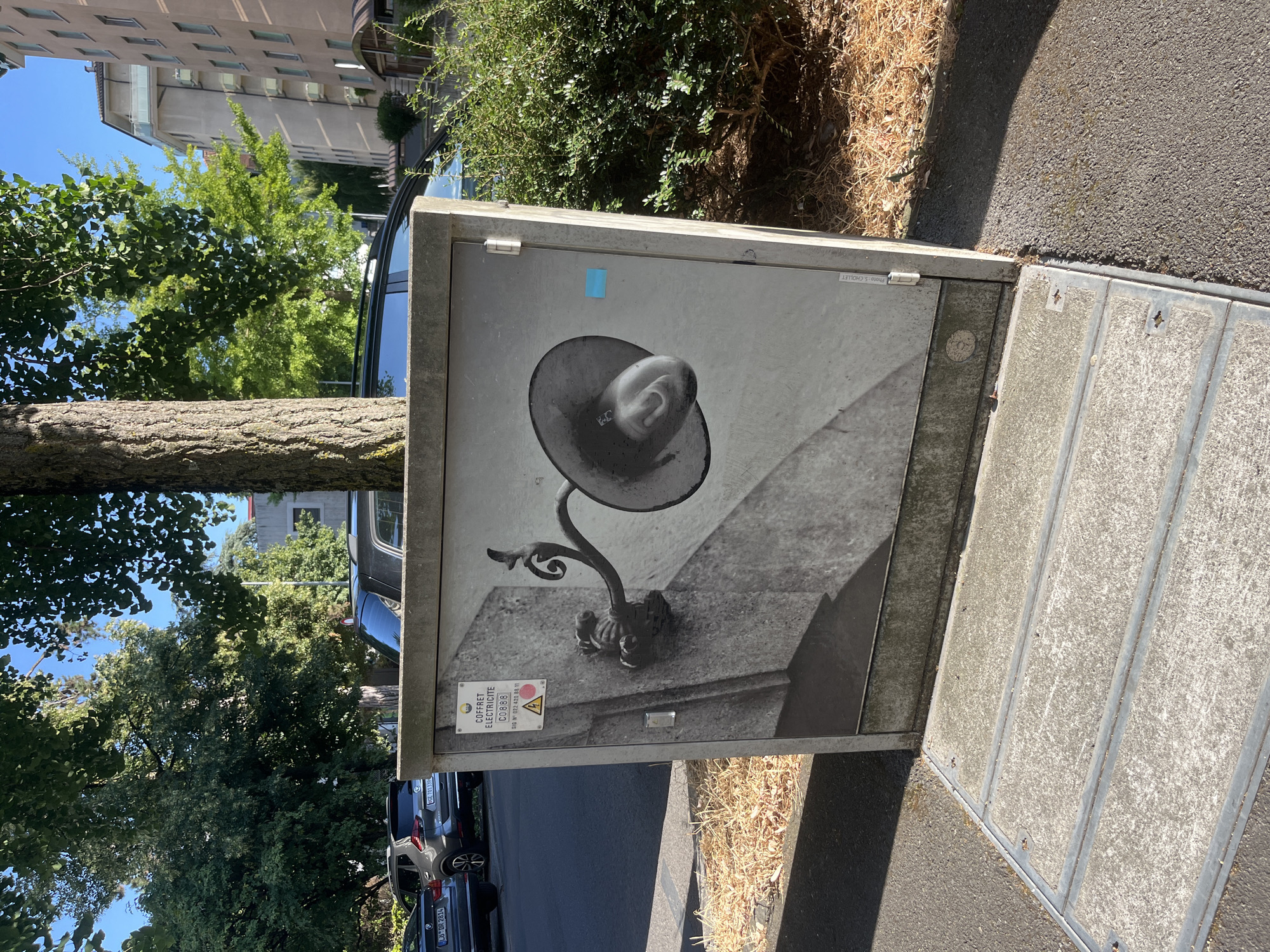 The image depicts a street scene in Geneva, Switzerland. In the foreground, a concrete utility box is adorned with a black and white mural of an old-fashioned street lamp. The mural covers the entire front face of the box. The box itself is gray and weathered, with a metal hinge visible on its right side. A small blue square sticker is affixed to the right side of the box. Above the mural, a sign is visible with text that reads "COFFRET ELECTRICITE". Below this, the numbers "C0888" are printed. A triangular warning sign with a red circle and a dot is also present. To the left of the utility box, a portion of a dark gray car is visible. Behind the utility box, a tree trunk with rough bark stands prominently, with lush green foliage overhead. Further in the background, several cars are parked along the street, and the facade of a tall, multi-story building with balconies can be seen. The ground is a mix of asphalt and dry grass. The scene is bathed in bright sunlight, casting distinct shadows.