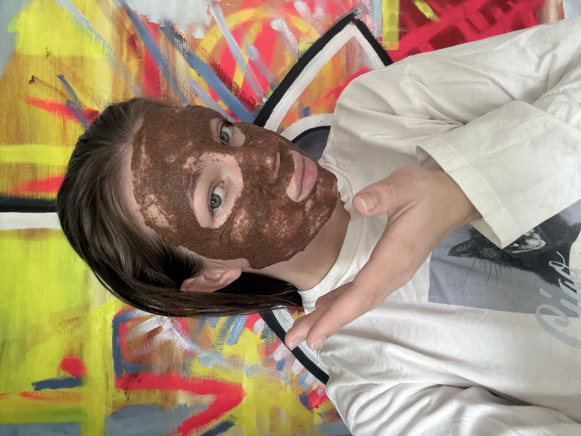 A person with a dark brown facial mask lies on a bed or similar surface. The person wears a white long-sleeved shirt featuring a graphic design. Their hair is dark, and their skin is fair except for the face mask. One hand is extended toward the camera. A colorful abstract painting hangs on the wall behind them. The location cannot be determined from the image.
