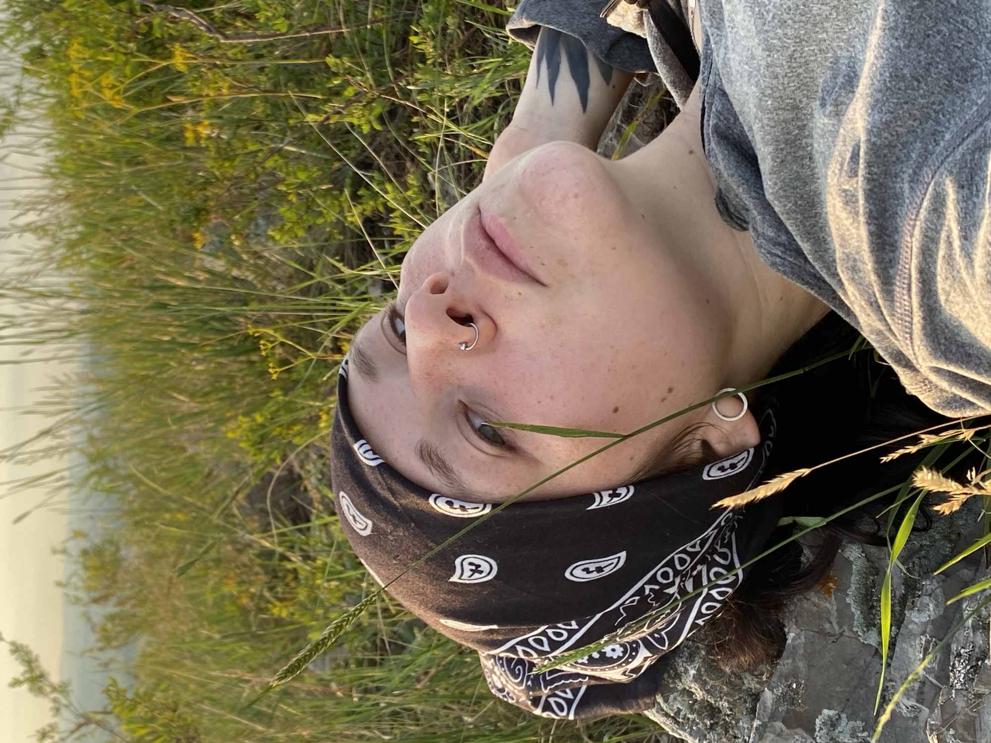 A person with a bandana wrapped around their head lies on a rock outdoors, surrounded by tall grass and vegetation. The person is looking upwards with their eyes open. They have a nose ring and an earring. A tattoo is visible on their arm. The background suggests a natural, possibly rural landscape with a hazy sky. The lighting indicates it may be late afternoon or early evening. There is no indication of the city of Kondurovka, Russia in the image or its description.