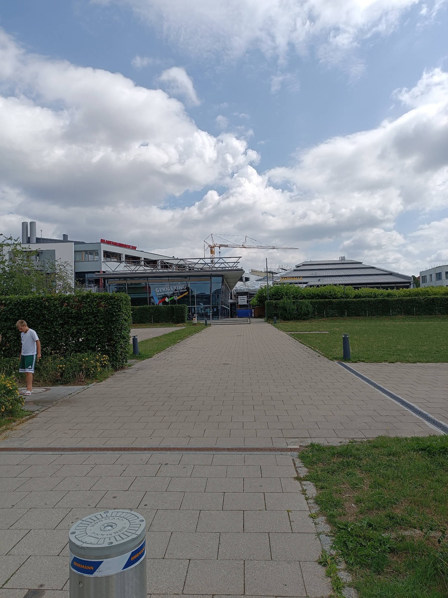 The image shows the exterior of the Stadthalle Germering, located in Germering, Germany. A young boy in a white t-shirt and green shorts stands to the left, looking down at some plants. In the background, the Stadthalle is visible, characterized by its distinctive layered roof structure. A large sign with the text "STADTHALLE" is mounted on the roof. Several cranes are also present, indicating ongoing construction or renovation work. The area features a paved walkway leading towards the building, bordered by green lawn and hedges. A cylindrical bollard with "HORMANN" branding is visible in the foreground. The sky is partly cloudy with a mix of white and gray clouds.