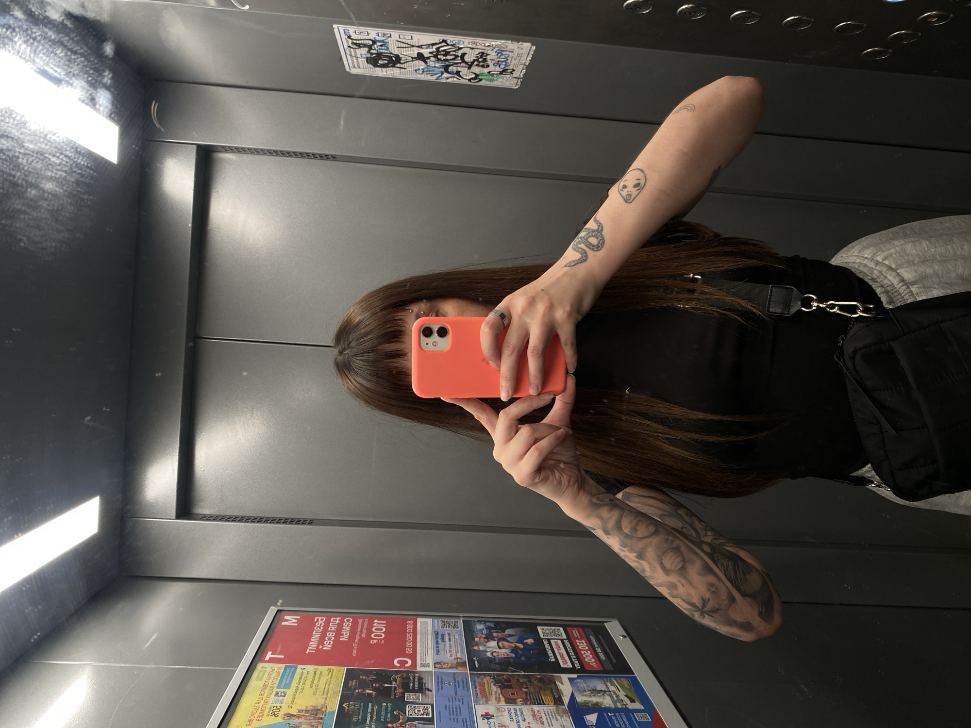 In an elevator in Votkinsk, Russia, a person with long brown hair and bangs, wearing a black shirt, grey sweatpants, and a black crossbody bag, takes a selfie. They are holding a bright orange smartphone in front of their face, obscuring it. The elevator's walls are dark grey metal, with a large poster on the left side featuring various images and text in Russian. On the right wall, there is graffiti. The elevator doors are closed, and a fluorescent light illuminates the space from above. The person has tattoos on their arms, including a snake design on their right forearm and abstract shapes on their left arm. A piercing is visible on their eyebrow.