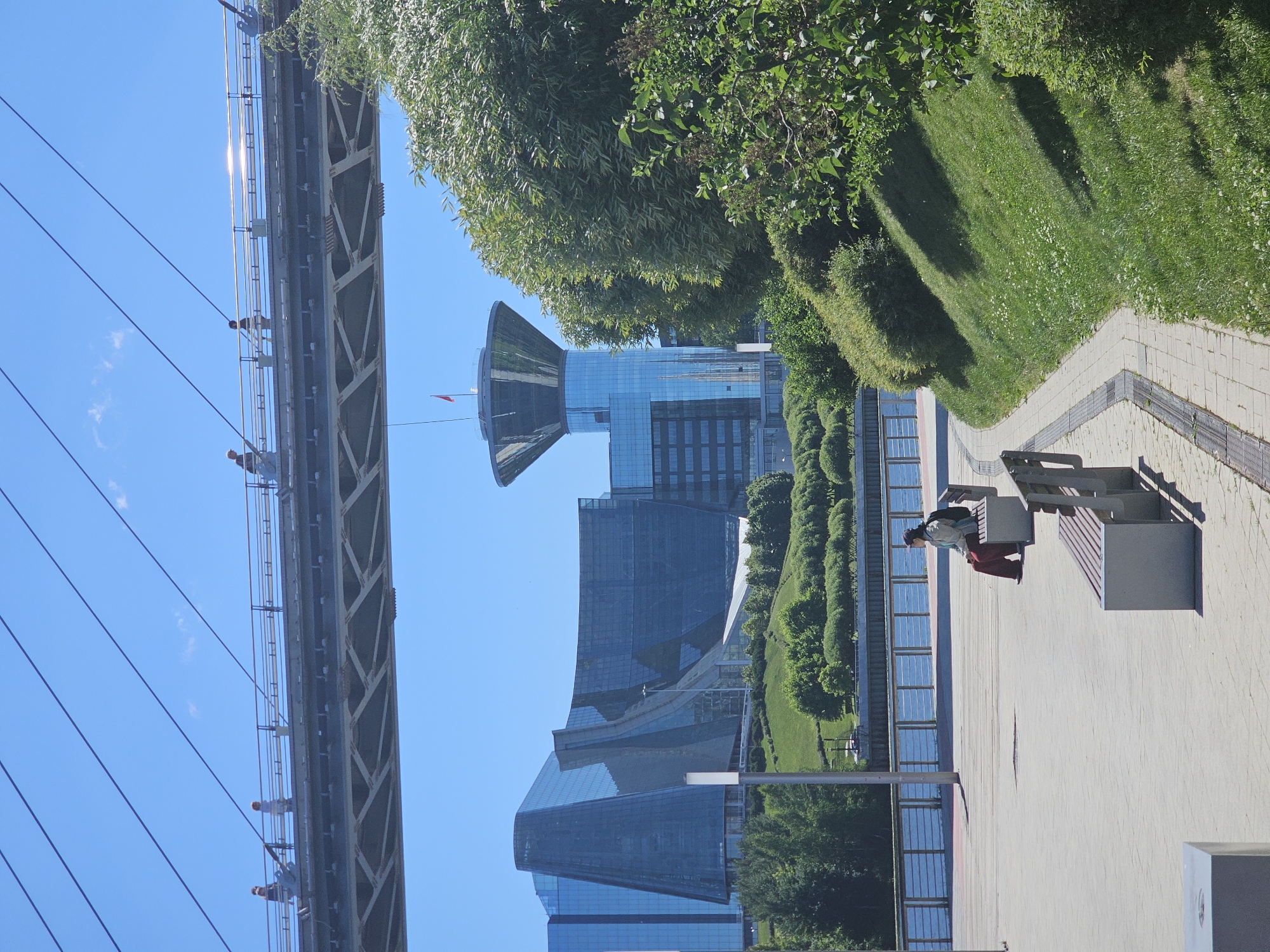 This is a full-shot, eye-level, outdoor photograph taken on a sunny day in Khimki, Russia, likely along the Moscow Canal. In the foreground, a paved walkway leads past empty park benches and a sloped grassy hill covered in green grass. A woman with dark hair, wearing a light green jacket and maroon pants, sits on a bench facing right, with a black backpack on the bench beside her. In the background, a suspension bridge with people walking on it spans across the frame. Beyond the bridge, modern, angular glass buildings rise, with one distinctive tower featuring a wide, flared top resembling a water tower. The buildings are nestled into a green, hilly landscape with mature trees. The sky is a clear, bright blue.