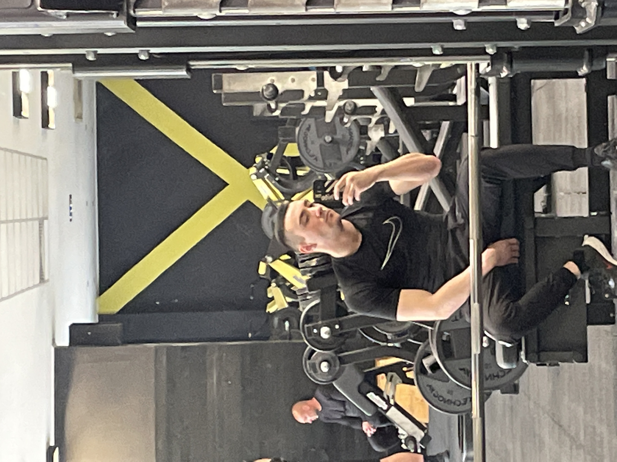 A man is taking a selfie while using a leg press machine in a gym. The gym has black walls with yellow stripes. Weight plates are loaded on the machine. Another person is visible in the background. The location is most likely Plovdiv, Bulgaria.