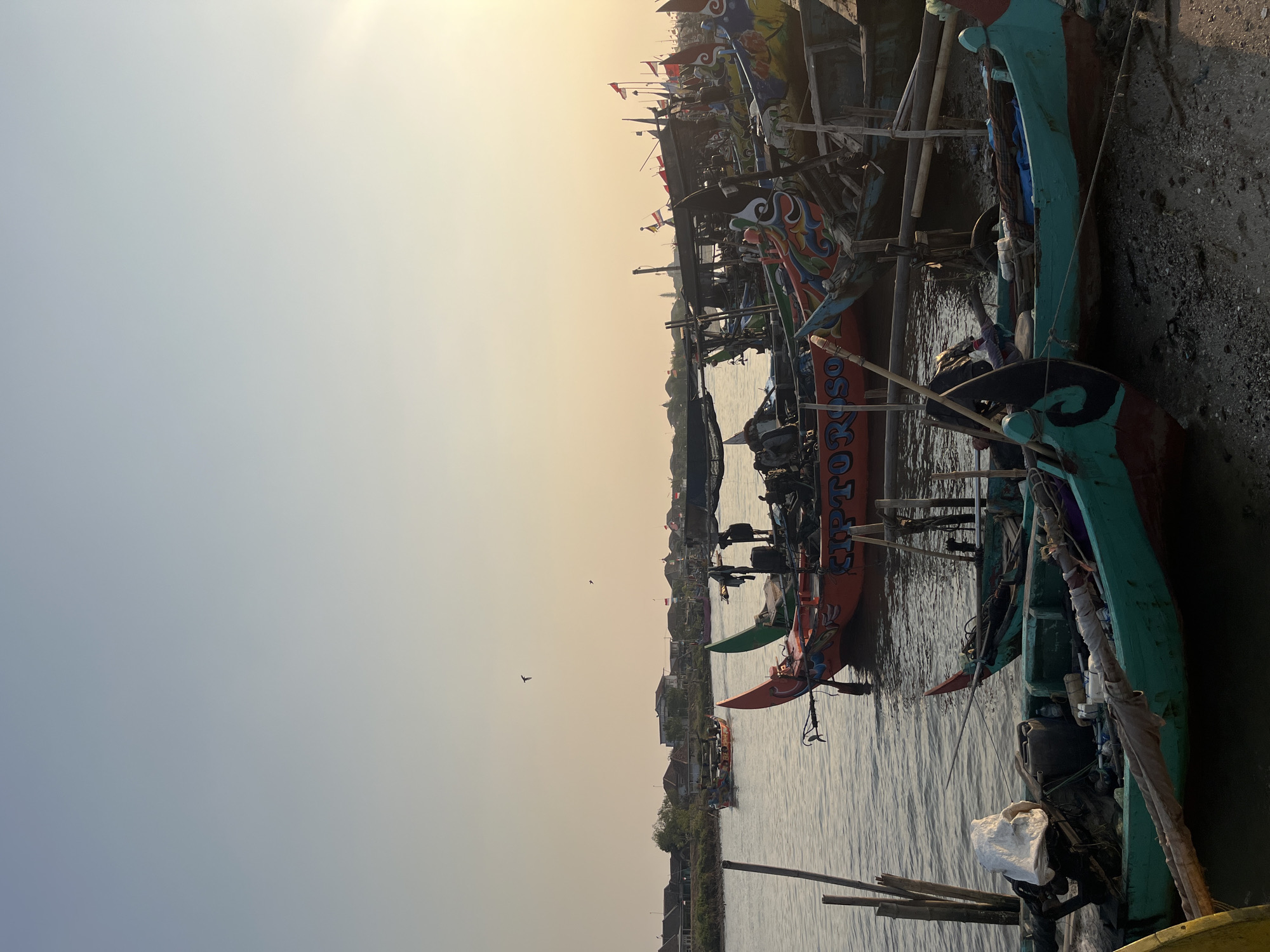 In Kedungkarang, Indonesia, a collection of colorful fishing boats are docked along a shoreline. The scene, captured at sunset, shows the sky a soft gradient of blue and yellow. Several boats are visible, their hulls painted in vibrant hues of orange, teal, and red, adorned with decorative patterns. Some boats are partially submerged in the calm water, while others rest on the sandy shore. The boats are laden with equipment, including nets, ropes, and containers, suggesting preparation for or return from fishing. In the distance, a town with houses and buildings lines the waterfront. A few birds are flying in the sky. The overall atmosphere is serene, indicative of a coastal community at the end of the day.