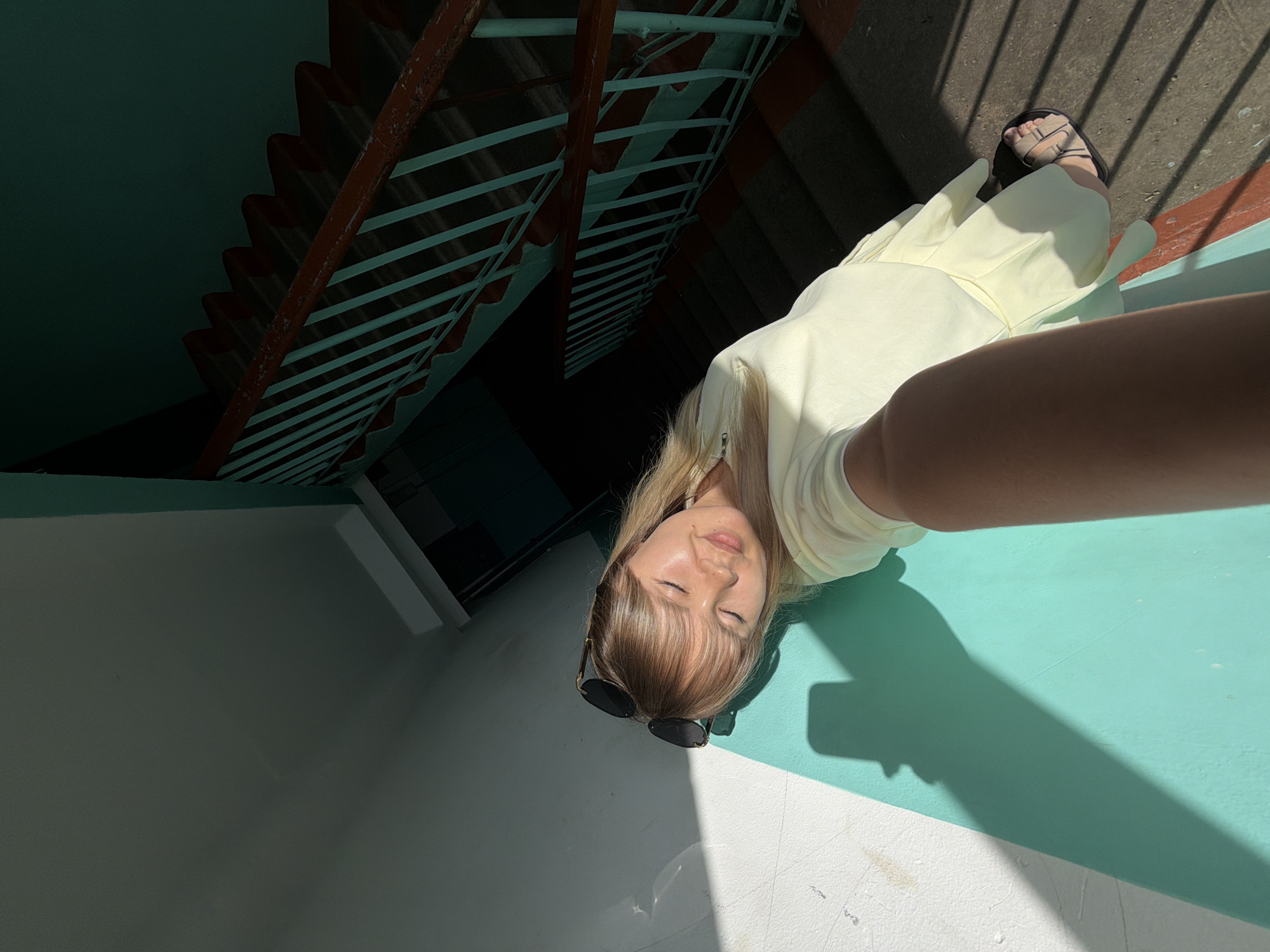 A woman with blonde hair and bangs is lying down outdoors, with her head resting on a teal-colored surface. She is wearing a light yellow dress and sandals. Sunglasses are perched on her nose. The scene is brightly lit by sunlight, casting strong shadows, suggesting it is daytime. In the background, a dark staircase with red and teal railings is visible. The overall impression is one of relaxation, possibly during a sunny day in Chaikovsky, Russia.