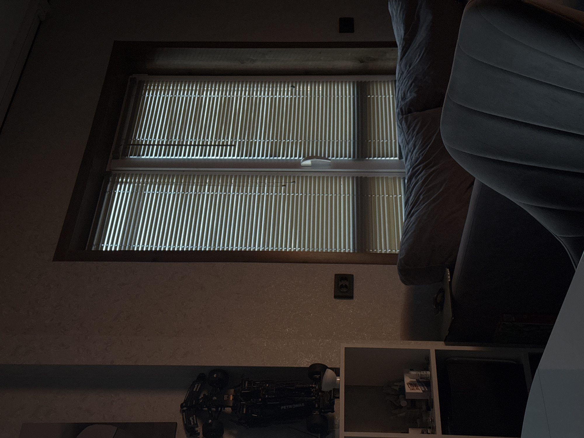 The image depicts an indoor scene, appearing to be a room such as a living area, with subdued lighting. The photograph is oriented 90 degrees clockwise from its natural perspective. When viewed correctly, a large window fitted with vertical blinds or slats occupies the upper left section. Bright natural light filters through the closed blinds, creating distinct vertical patterns of illumination on the interior side.
To the right, a dark-colored sofa or armchair with multiple upholstered segments is partially visible. The room features light-colored, textured walls. Below the window, resting on a dark surface, is a black model car or intricate toy vehicle. The word "PETRONAS" is clearly visible in white lettering on its side. An electrical wall outlet is positioned on the wall below the window. Further to the right, a white shelving unit with open cubbies is visible. One cubby contains various small objects, including a white box with some red, illegible text.
The scene suggests daytime due to the bright light from the window, though the interior remains dim, possibly due to the blinds and the room's orientation or time of day. No human activity is present. The objects indicate a personal space or hobby area. The image does not contain specific visual cues to its reported location in Лобаново, Russia.