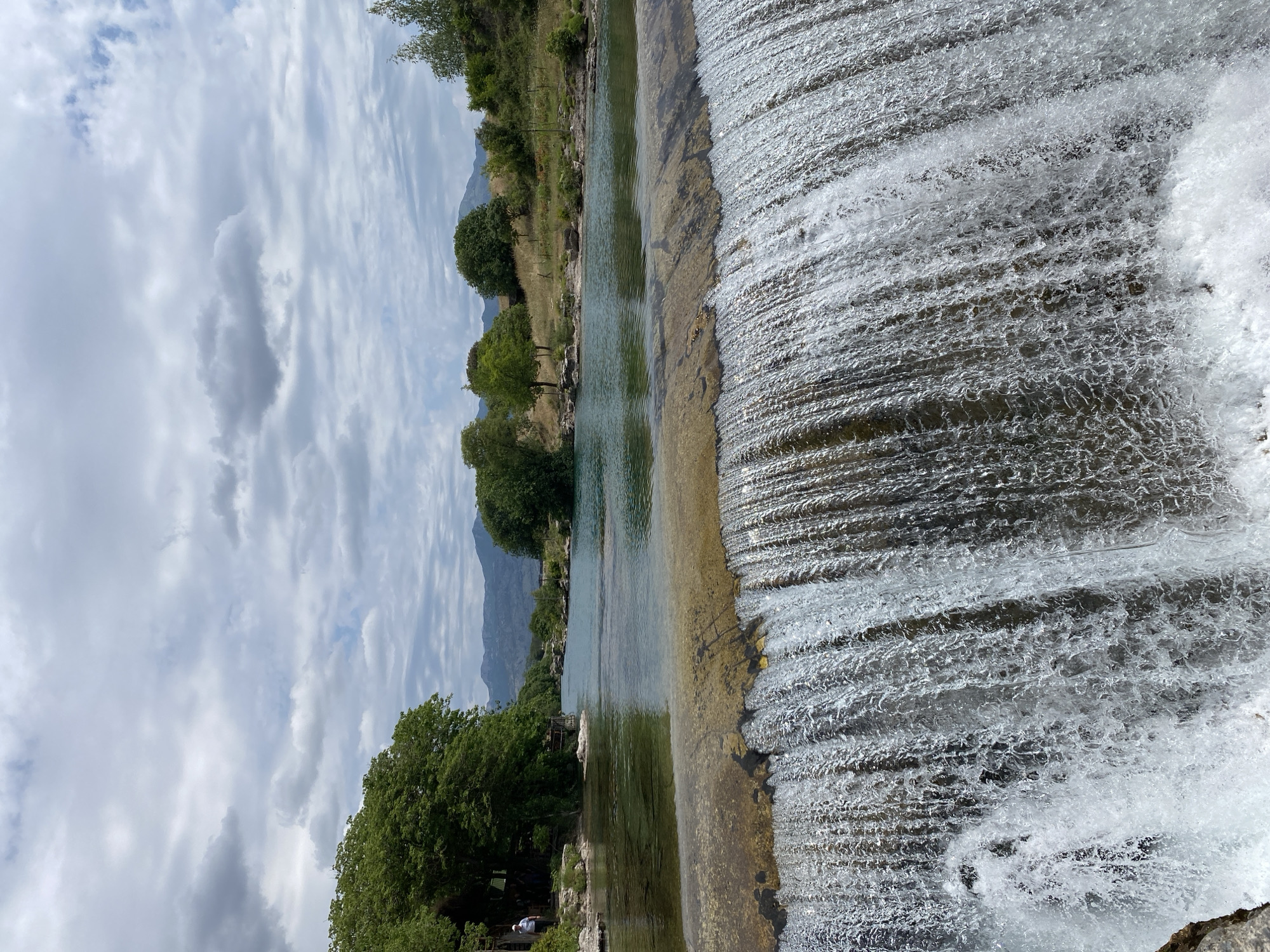 This image captures a serene natural landscape featuring a wide river with a cascading waterfall. The scene is dominated by clear, flowing water that appears shallow enough in places to reveal a rocky, light-colored riverbed. On the right side, the water spills over a wide, low barrier, creating a sheet-like waterfall that extends horizontally across much of the frame, with visible frothing and splashing at its base.
Lush green trees and vegetation line both banks of the river, indicating a verdant environment. A variety of trees with dense canopies populate the mid-ground and foreground, especially along the left bank. In the distance, mountains rise softly against a partly cloudy sky, which features expansive grey and white clouds with occasional patches of brighter sky.
In the lower left portion of the image, two small figures are discernible near the river's edge, nestled among the trees. One figure is wearing a white top, suggesting people are casually enjoying the natural setting.
The time of day appears to be daytime, possibly under an overcast or partly sunny sky, creating a soft, diffused light over the scene. The weather is cloudy but bright, contributing to a tranquil atmosphere. The setting is indicative of a picturesque river environment, consistent with the provided location context of Podgorica, Montenegro. No visible text is present in the image.