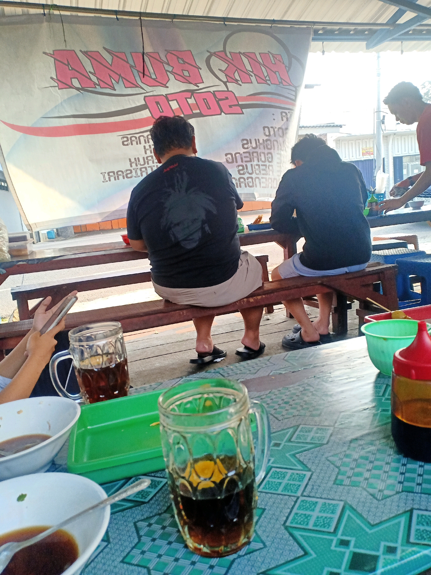 The image depicts an outdoor dining area, likely a warung or roadside eatery, with a banner overhead that has Indonesian text. The banner reads "AMUB OTO" and has other text that is difficult to read. Three people are visible. Two men are seated on a wooden bench, facing away from the camera. One man wears a black t-shirt with a graphic design on the back and light-colored shorts. The other man wears a dark blue long-sleeved shirt and light blue shorts. A third person, partially visible on the left, holds a smartphone. In the foreground, a table is covered with a patterned tablecloth. On the table are two glasses of iced tea, two white bowls containing some liquid and a spoon, a green plastic tray, and a red-capped bottle. The background shows a building and some utilities, and the overall setting suggests a warm climate. This scene is consistent with a warung located in Jatisrono, Indonesia, a district known for its roadside eateries and local businesses.