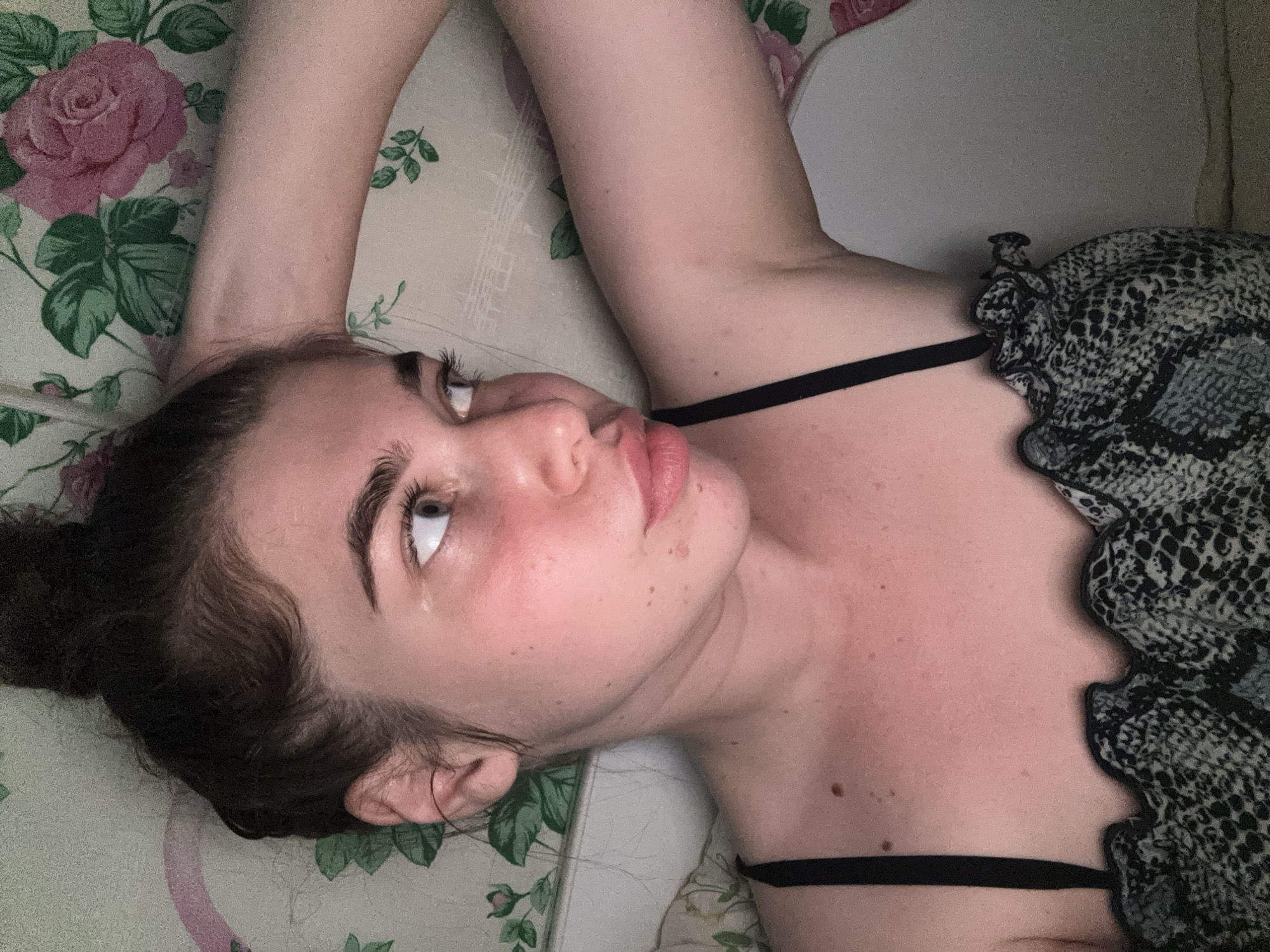 A young woman with her hair in a bun is lying on her side, propped up on her arm. She is wearing a black spaghetti strap top with a ruffled neckline that has a snake-skin pattern. The background is a floral patterned fabric, possibly a pillowcase or sheet, with large pink roses and green leaves. The woman's face is turned to her right, and she appears to be looking away from the camera. Her skin is flushed, particularly on her cheeks and neck. The lighting is soft and appears to be coming from the side. There are no visible landmarks or indications of the city of Мещёры, Russia, in the image.