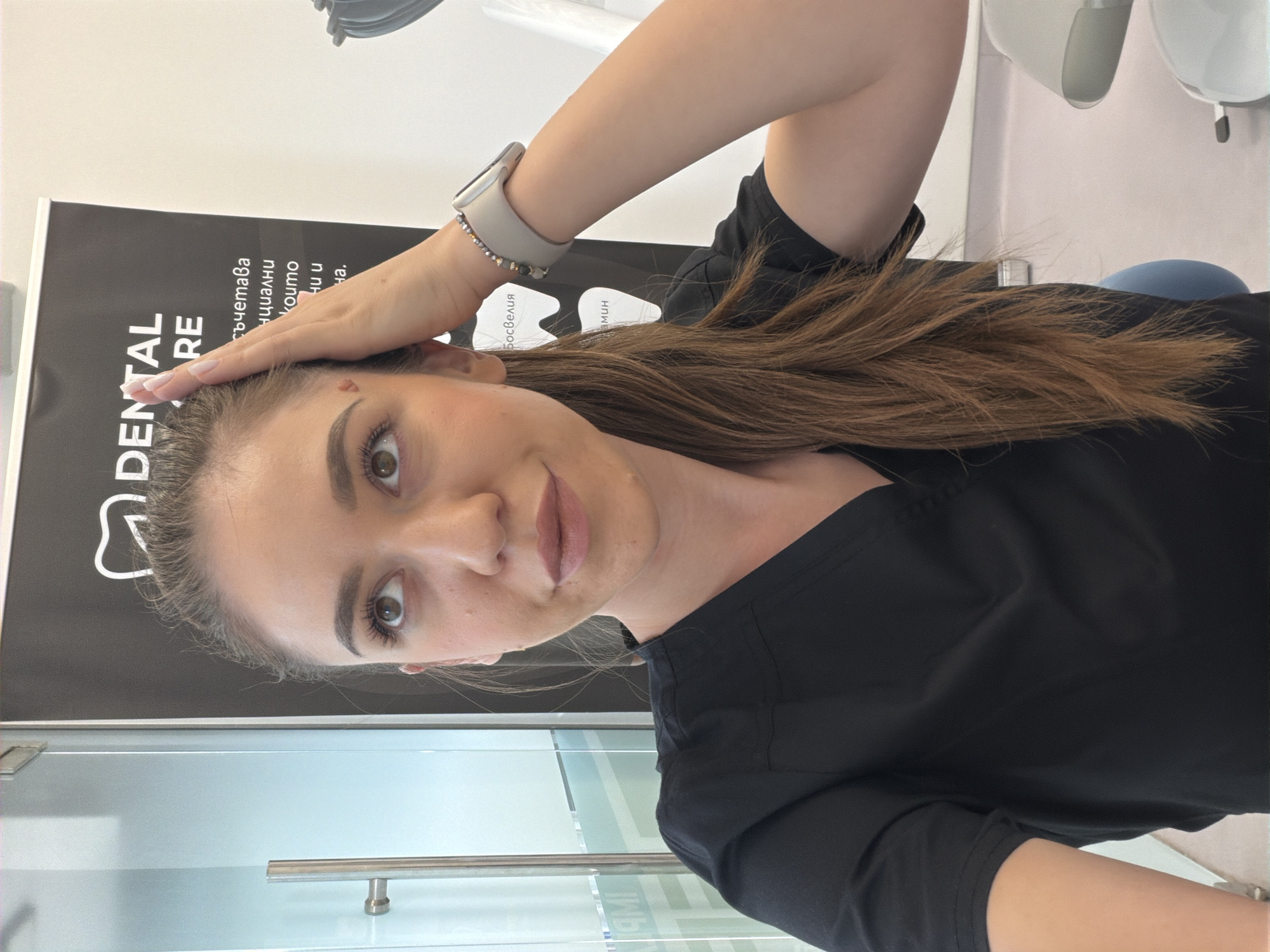 A young woman with long, brown hair is pictured indoors, seemingly in a dental office. She is wearing a black scrub top. She has her right hand on her forehead, her arm bent at the elbow. Her hair falls over her shoulder. She is wearing a light-colored smartwatch on her left wrist, adorned with small, dark beads.
Behind her, to her left, is a black banner with white text. The text appears to be in Bulgarian. The words "DENTAL CARE" are clearly visible in large white letters on the banner. Beneath this, smaller text is also present.
The setting appears to be a clean, modern environment, possibly a waiting area or treatment room in a dental clinic. The lighting suggests it is daytime. The woman's expression is neutral, as she looks directly at the camera. The image is oriented sideways, with the woman's head to the left.