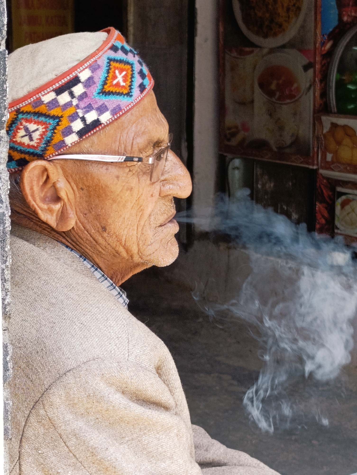 This is a medium-shot, profile view of an elderly South Asian man smoking a cigarette. He is wearing a traditional wool cap with a colorful woven band featuring geometric patterns in purple, orange, blue, and white, including diamond shapes with an 'X' motif. The man has weathered skin, glasses with thin wire frames, and his mouth is slightly open, exhaling a plume of white smoke that drifts to the right. He is wearing a beige woven garment with a checkered collar peeking out from underneath.
The background appears to be an outdoor setting, possibly a shop or stall. Behind the man, there are framed images of food items displayed on a wall. One image shows what looks like a bowl of rice and another a dish with red sauce. The overall lighting suggests it is daytime. The texture of the man's clothing and cap, along with the smoke, are clearly visible. The setting is likely a public area given the display of food. The man's expression appears neutral, focused on the act of smoking. No specific text is clearly legible, although there are faint signs in the background which are out of focus. The location context points to Vashisht, India, a place known for its natural hot springs and Hindu temples.
