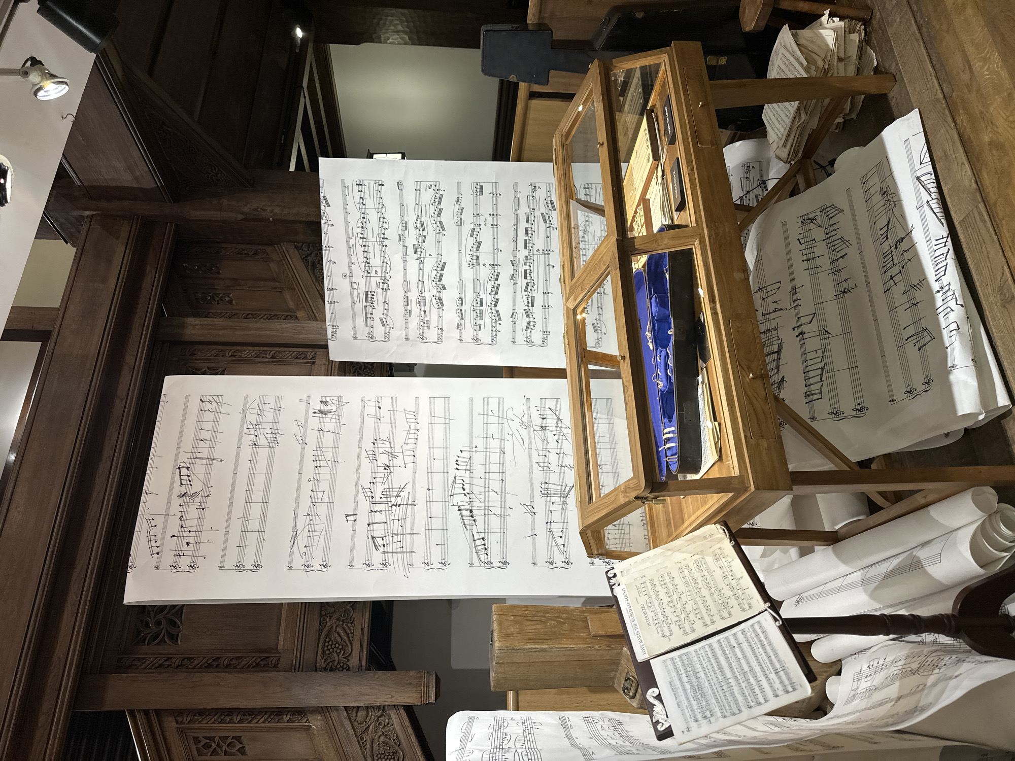 The image depicts an interior scene, possibly an exhibit or display, featuring musical scores and related items. Two large sheets of music are displayed vertically, appearing to be hung or supported in the center of the frame. To the right is a wooden display case containing an object wrapped in blue fabric. Below the case, an open book of sheet music rests on a pile of other music sheets and rolled-up documents. The surrounding architecture includes ornate dark wooden paneling, suggesting a historic or traditional setting. The lighting appears to be a combination of natural and artificial light, with a spotlight visible in the upper left corner. Without additional information, the specific location within London is impossible to determine.
