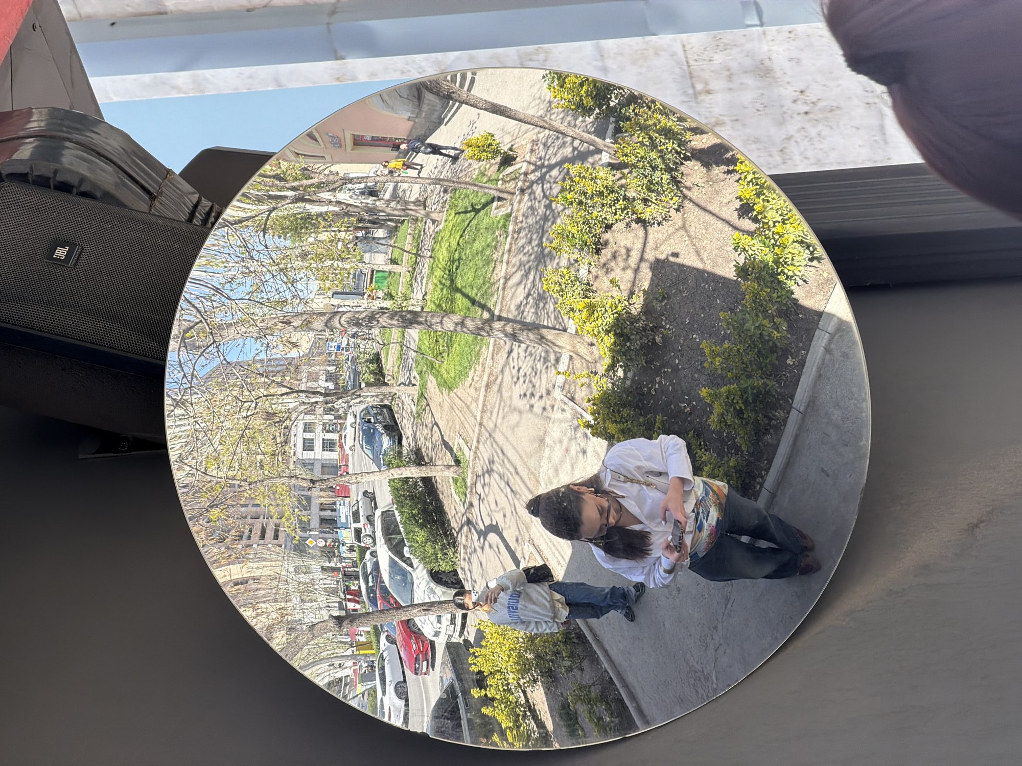 The image displays a circular mirror reflecting an outdoor urban scene, captured from an elevated perspective. The mirror rests on a flat, dark gray surface. To the left, a black JBL speaker is partially visible, angled downwards, with the "JBL" logo clearly displayed. Above the mirror and speaker, a clear blue sky is seen, while a dark, blurry object is present in the upper right corner.
The reflection within the mirror shows a street scene in Yerevan, Armenia, rotated approximately 90 degrees clockwise. Two people are visible. One person, closer to the viewer and in the foreground of the reflection, wears a white shirt and dark pants, holding a phone. Further into the reflected scene, another person is seen on a sidewalk, wearing a light gray hoodie with partially legible text (possibly "inventor" or "innova...") and blue jeans, also appearing to hold a phone.
The outdoor environment features a street lined with various parked cars, including white, red, and dark-colored vehicles. A sidewalk runs alongside a grassy area dotted with trees, some with bare branches suggesting early spring, and others with light green foliage. Small bushes with vibrant yellow flowers are interspersed throughout. In the background, multi-story buildings are visible, including one with a reddish-pink facade. Bright sunlight casts distinct shadows from the trees onto the ground, indicating a clear and sunny day, likely during mid-morning or afternoon. No specific emotions are discernible.