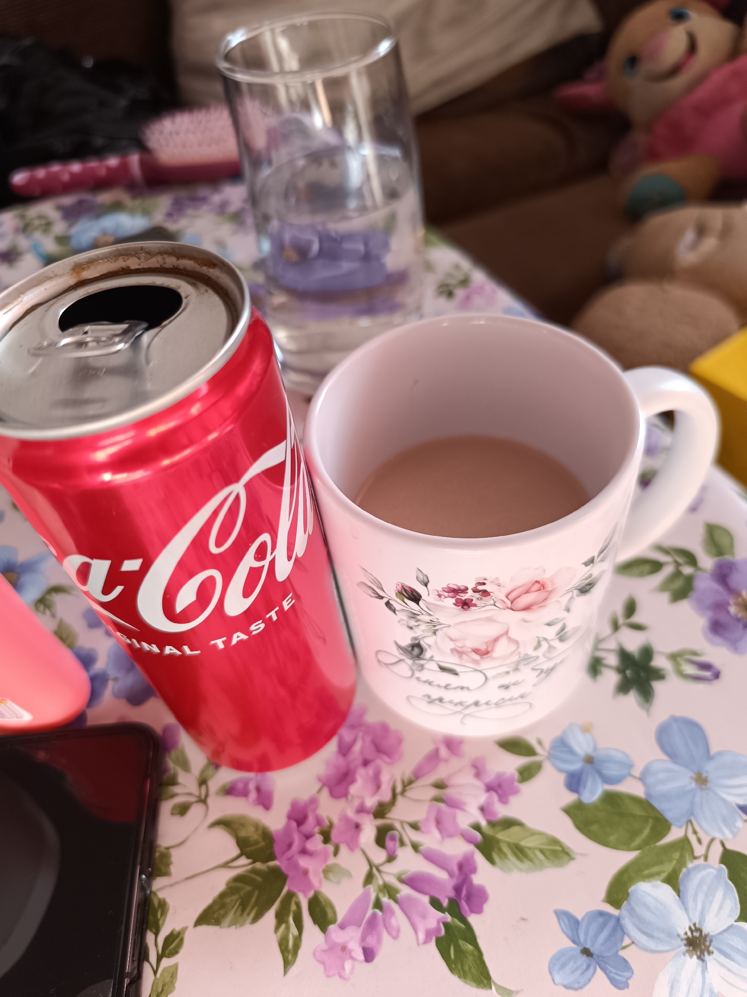 A close-up shot shows a can of Coca-Cola, a white floral-patterned mug filled with a light brown liquid, and a partially filled clear glass of water on a table covered in a floral-patterned cloth. A pink hairbrush and a black smartphone are also on the table. In the background, a blurry, brown sofa and plush toys can be seen. The location is Unknown, Unknown. No people are visible in the image.