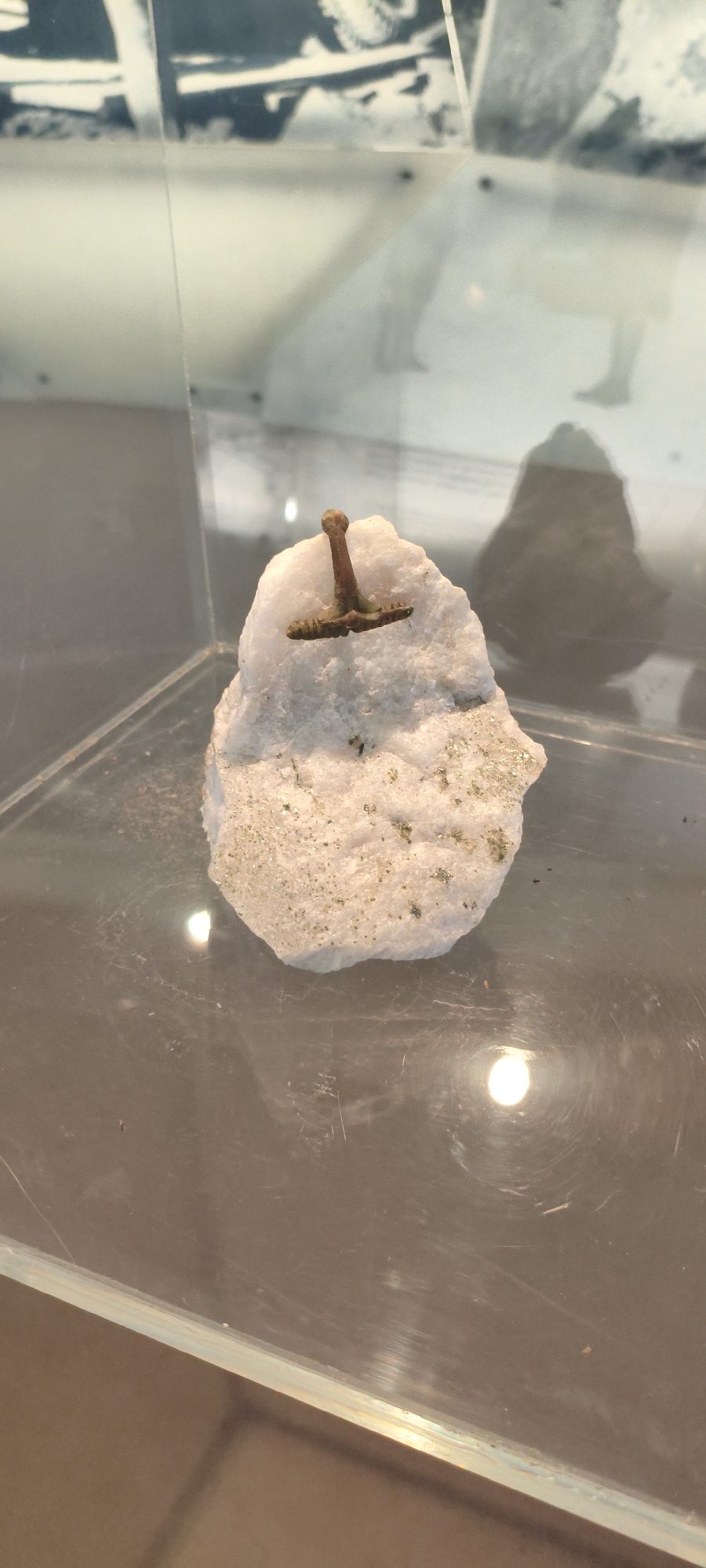 The image displays a small, ancient metal artifact, resembling a miniature hammer or a stylized T-shape, embedded in a chunk of white, crystalline rock. The artifact appears to be made of a dark, possibly bronze or iron, material and shows signs of age and patina. The rock itself has small, glittering flecks, possibly pyrite or another mineral inclusion. This display is enclosed within a glass case, suggesting it is an exhibit in a museum or exhibition. The background is blurred, showing reflections on the glass and what appears to be a black and white historical photograph or display panel above. The lighting creates reflections on the glass surface and the artifact. This artifact is located in the Sölden Ice AGE museum in Sölden, Austria.