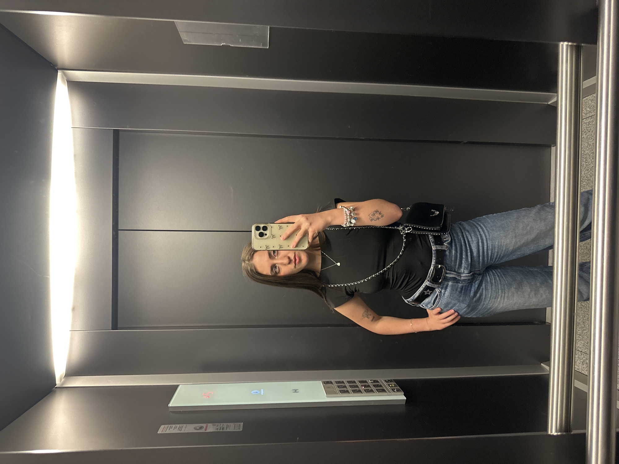 A young woman takes a selfie in an elevator. She is wearing a black shirt, blue jeans, a black purse, and jewelry. The elevator has a metal panel with buttons. The elevator is located in Huixquilucan de Degollado, Mexico.