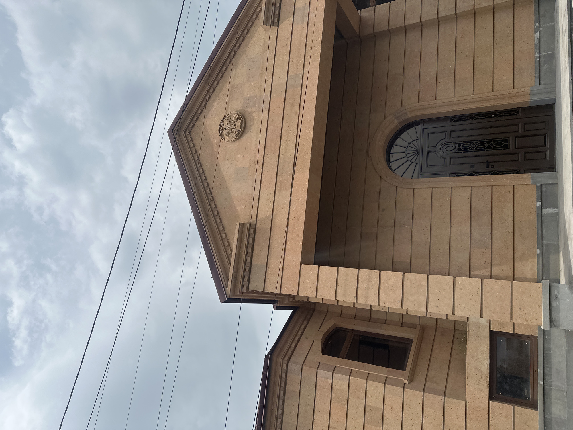 The image shows a close-up of the facade of a stone building in Yerevan, Armenia. The building has a gabled roof and is constructed of light brown stone blocks. There is a circular emblem on the gable. The front door is visible, with a rounded arch above it. There are two small windows on the lower level. Power lines are visible in the background against a cloudy sky. The overall impression is of a well-maintained building with a traditional architectural style.
