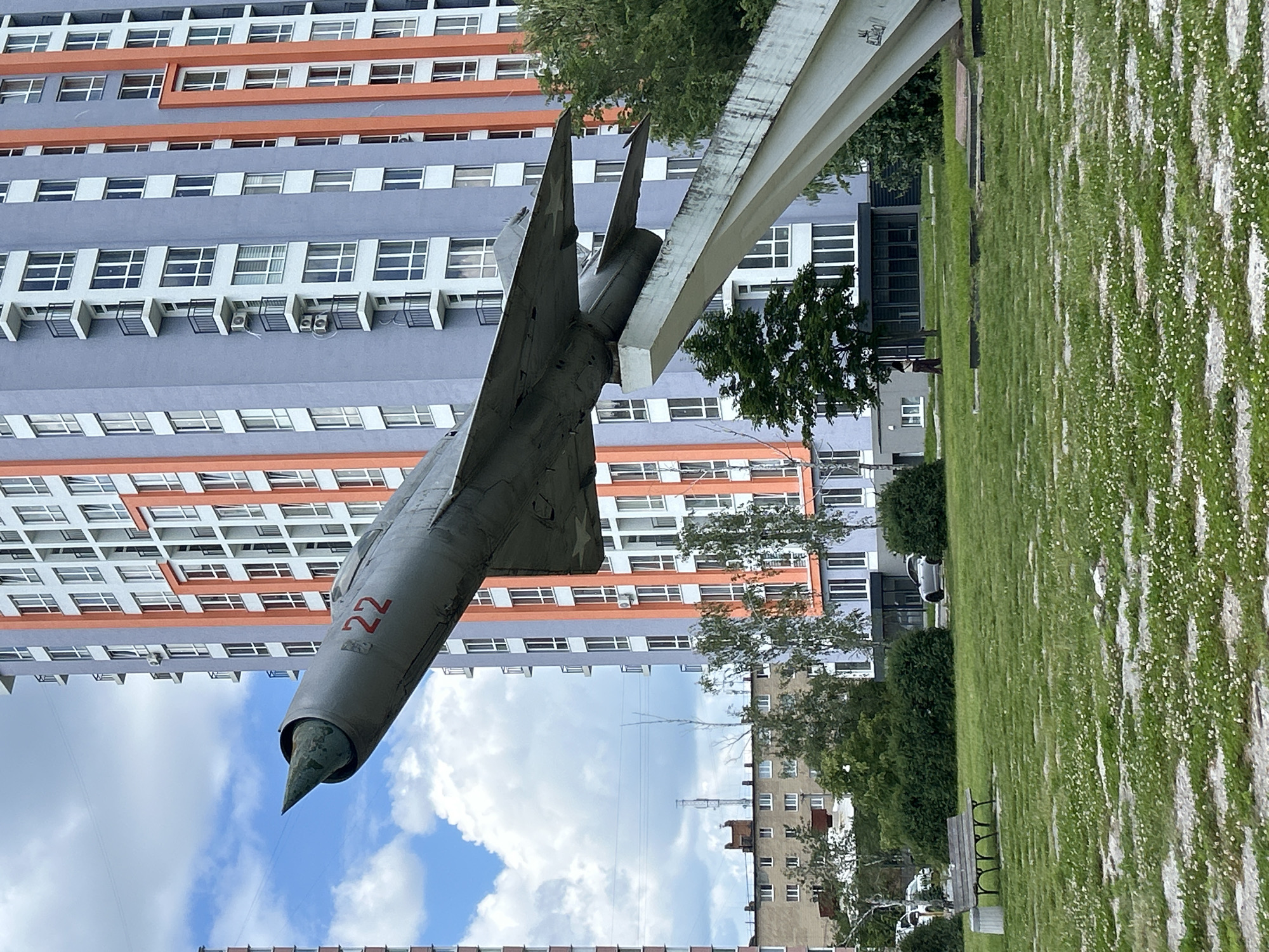 An old, gray fighter jet, with the number "22" painted in red on its fuselage, is mounted on a concrete pedestal in an urban park setting. The jet is angled upwards and to the left, its nose pointing towards a bright blue sky with scattered white clouds. Behind the jet, a modern apartment building with a striking blue and orange facade stands tall. The building features numerous windows and balconies. In the foreground, a neatly mown green lawn is interspersed with patches of gravel and small white flowers. A wooden park bench and a trash bin are visible on the left side of the image. Further in the background, another, older apartment building with a beige facade can be seen, along with a few trees. A person is standing on the right side of the image, near the base of the jet's pedestal. The scene appears to be located in Bila Tserkva, Ukraine, given the architectural style of the buildings.