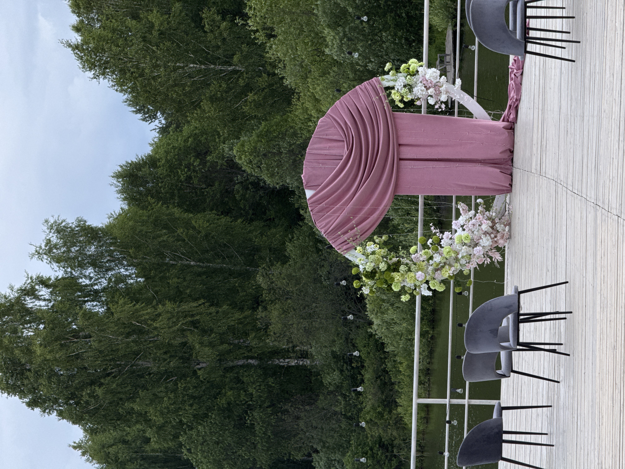 An outdoor wedding setup is depicted on a wooden platform near a body of water, likely a lake, with a dense forest bordering the opposite bank. The focal point is a circular wedding arch draped with a dusty rose fabric that cascades to the floor. Floral arrangements in shades of white and green adorn the arch. Several dark grey upholstered chairs with black legs are arranged on the platform facing the arch, suggesting a seating area for guests. String lights are hung along a white railing that separates the platform from the water. The overall atmosphere is serene and natural, with the overcast sky indicating a potentially temperate climate. The location is identified as Votkinsk, Russia. There are no people visible in the scene.