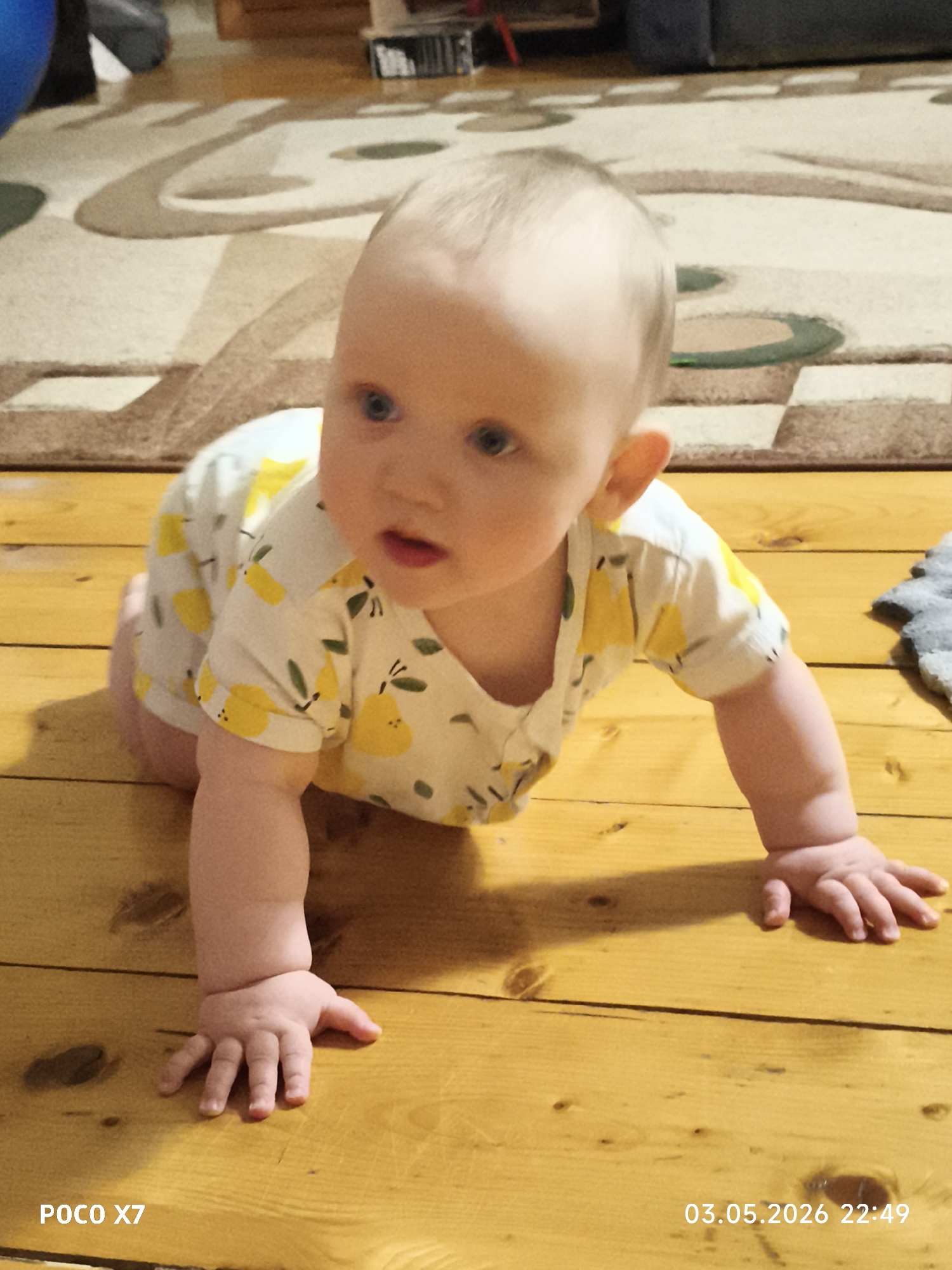 A baby crawls on a wooden floor, facing forward. The baby wears a white and yellow pear-patterned onesie. A patterned rug is visible in the background. The image contains a POCO X7 watermark and timestamp "03.05.2026 22:49". The location is unknown, although the timestamp is consistent with a date.