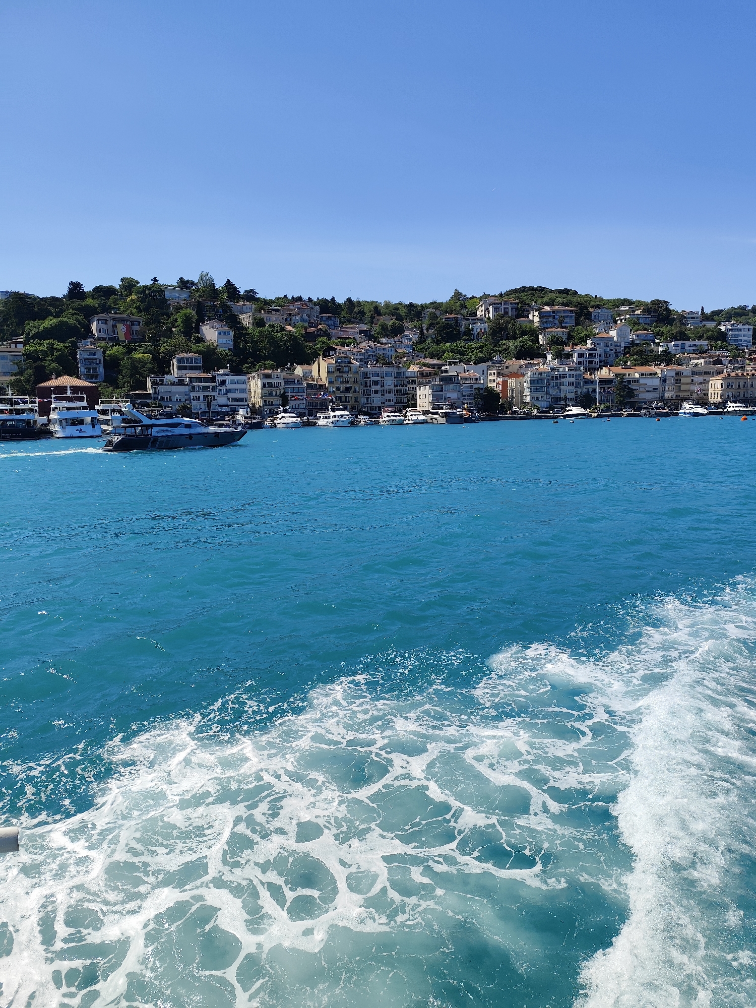 The image depicts a waterfront scene in Beşiktaş, Turkey, from the perspective of a boat moving through the water. The foreground features the foamy wake of the boat in the turquoise water of the Bosphorus strait. In the middle ground, several yachts and boats are visible along the shoreline. A large cluster of buildings, mostly white or light-colored, covers the hillside leading up from the water. Green trees punctuate the architectural landscape. The sky is clear and bright blue.