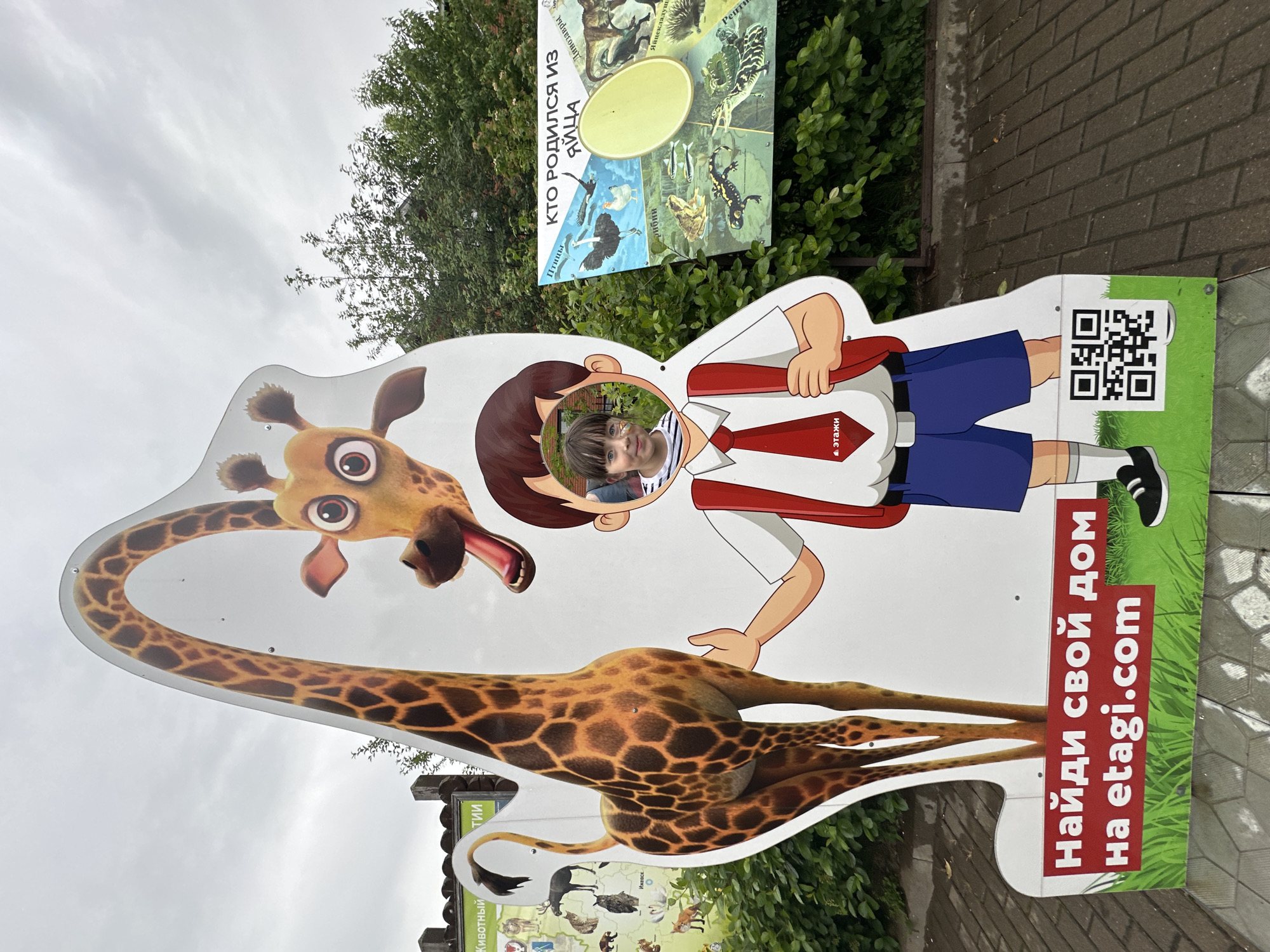 The image depicts a cutout stand with a cartoon giraffe and a cartoon boy, designed for visitors to pose for photos. A young boy with face paint is peeking through a hole in the cutout, his head and shoulders visible. He is wearing a striped shirt and a red tie with a logo. Behind the cutout, there are trees and foliage, suggesting an outdoor location. To the right of the cutout is a sign with pictures of animals that hatch from eggs, with Russian text indicating "Who was born from an egg." To the left of the cutout is another sign with images of animals and Russian text. At the bottom of the cutout, there is a green section with a red rectangle containing white Russian text that reads "Find your home on etagi.com" and a QR code. The ground appears to be paved with cobblestones. The overall scene suggests a public attraction, likely a zoo or an amusement park, within the city of Izhevsk, Russia.