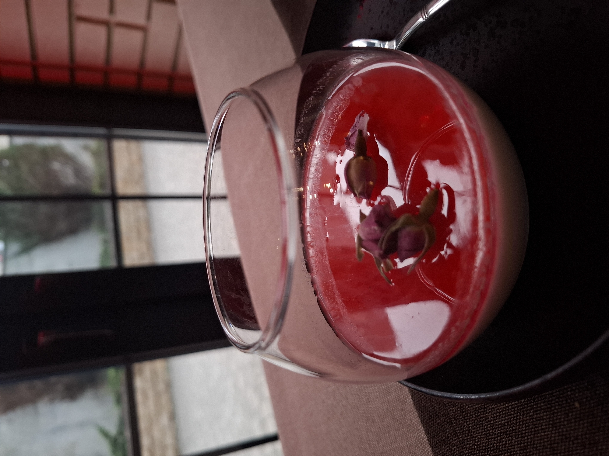 A glass of dessert sits on a black plate, set on a brown placemat. The dessert consists of a layer of pale cream at the bottom and a layer of translucent red liquid on top, garnished with two small flower buds. The dessert rests near a window that looks out onto a street scene, potentially in Zadonsk, Russia, given the prompt’s specification. The building’s interior has a red and brown color scheme. A silver spoon is placed near the glass.