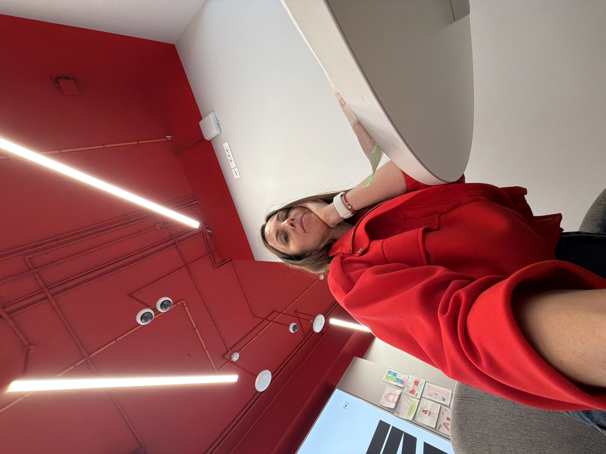 A person wearing a red jacket is in what appears to be a modern interior space with a red ceiling and white walls. Long, bright lights illuminate the red ceiling, and surveillance cameras are visible. The person has brown hair and is wearing a white wristband or smartwatch. A white object is positioned near them, possibly a computer screen or monitor. Children's drawings are displayed on a wall near a monitor with black elements visible. The location is not confirmed to be in Birobidzhan, Russia, as there are no elements confirming the location in the picture.