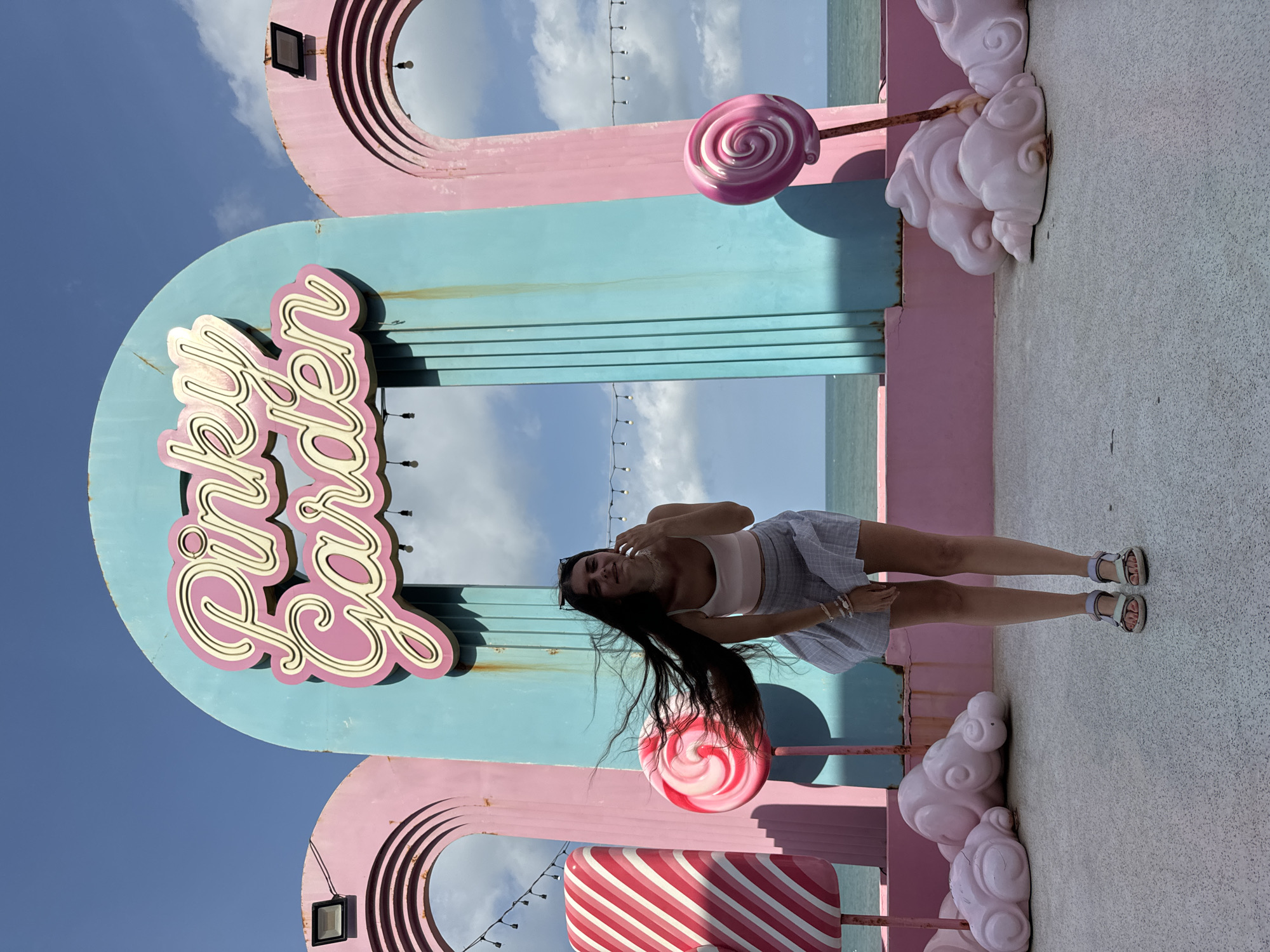 A young woman stands in front of an arched structure with "Pinky Garden" written on it in neon lights. The structure is painted in pastel pink and teal colors. The woman has long dark hair and is wearing a light pink tank top and a blue and white checkered skirt. She is wearing sandals and has several bracelets on her wrists. In the foreground, there are pink cloud-shaped sculptures and large lollipop decorations. The background shows the ocean and a bright blue sky with scattered clouds. The scene appears to be an outdoor recreational area or a themed attraction, possibly a tourist spot in Tien Thanh Ward, Vietnam, given the presence of the ocean and the overall aesthetic.