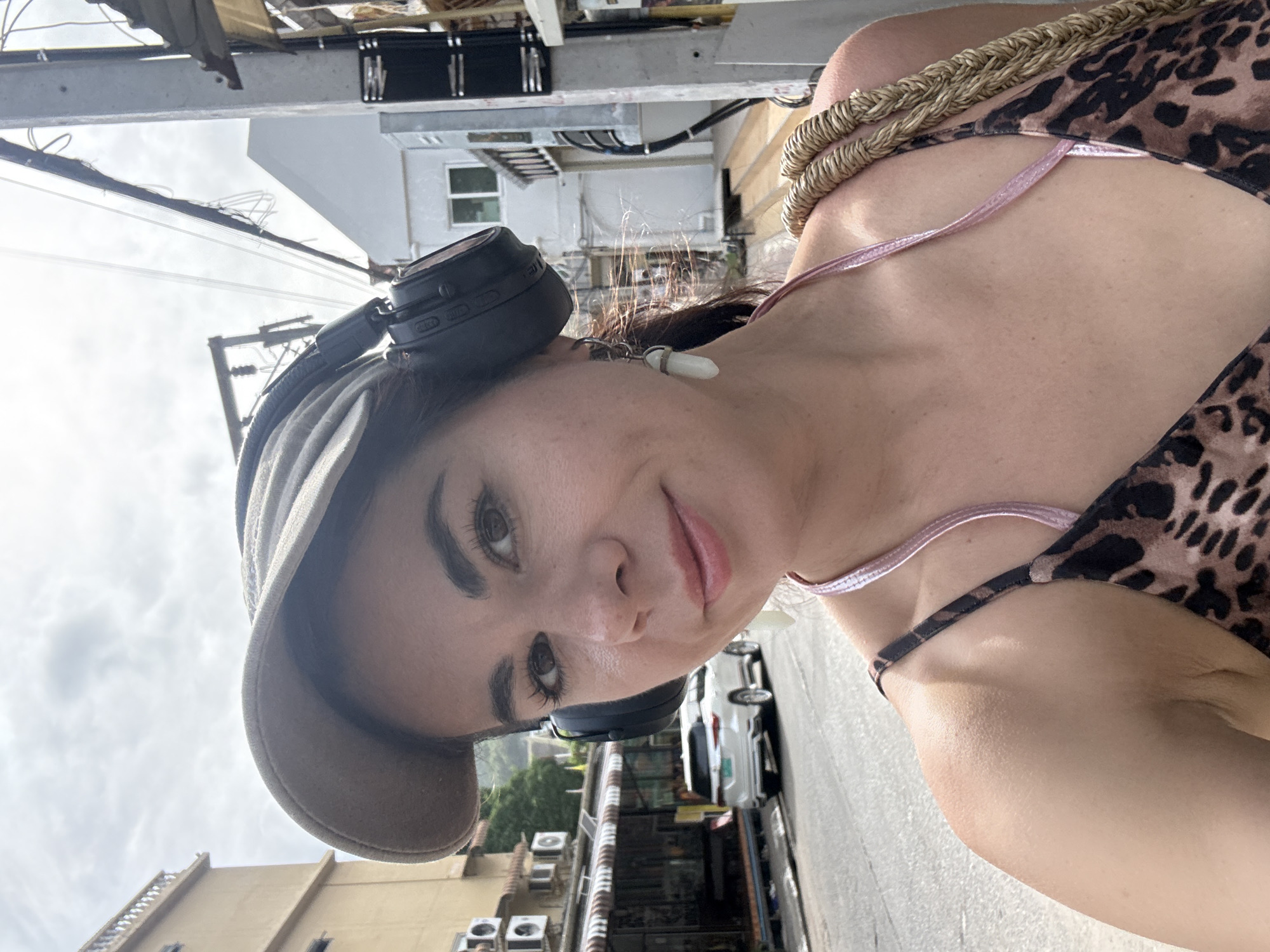 A woman stands on a street in Karon, Thailand, taking a selfie. She is wearing a baseball cap, headphones, and a leopard-print top with thin pink straps. She has light skin and brown eyes, and her hair is dark brown. She is also wearing long, light-colored earrings. In the background, a white SUV is parked on the side of the road, and buildings with awnings line the street. The sky is overcast.